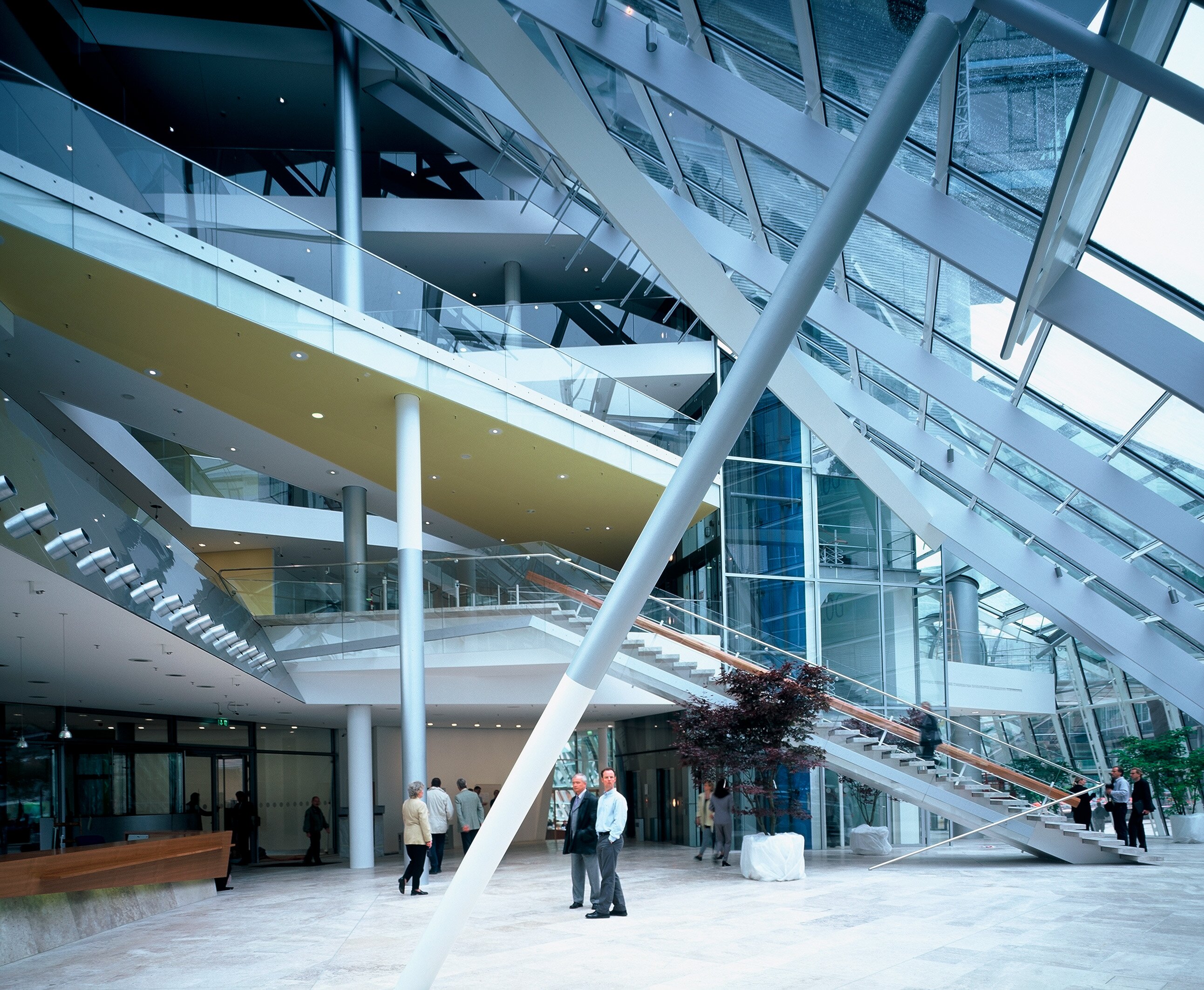 Spacious modern atrium with glass walls, large diagonal beams, stairs, and people walking and standing on the ground floor.