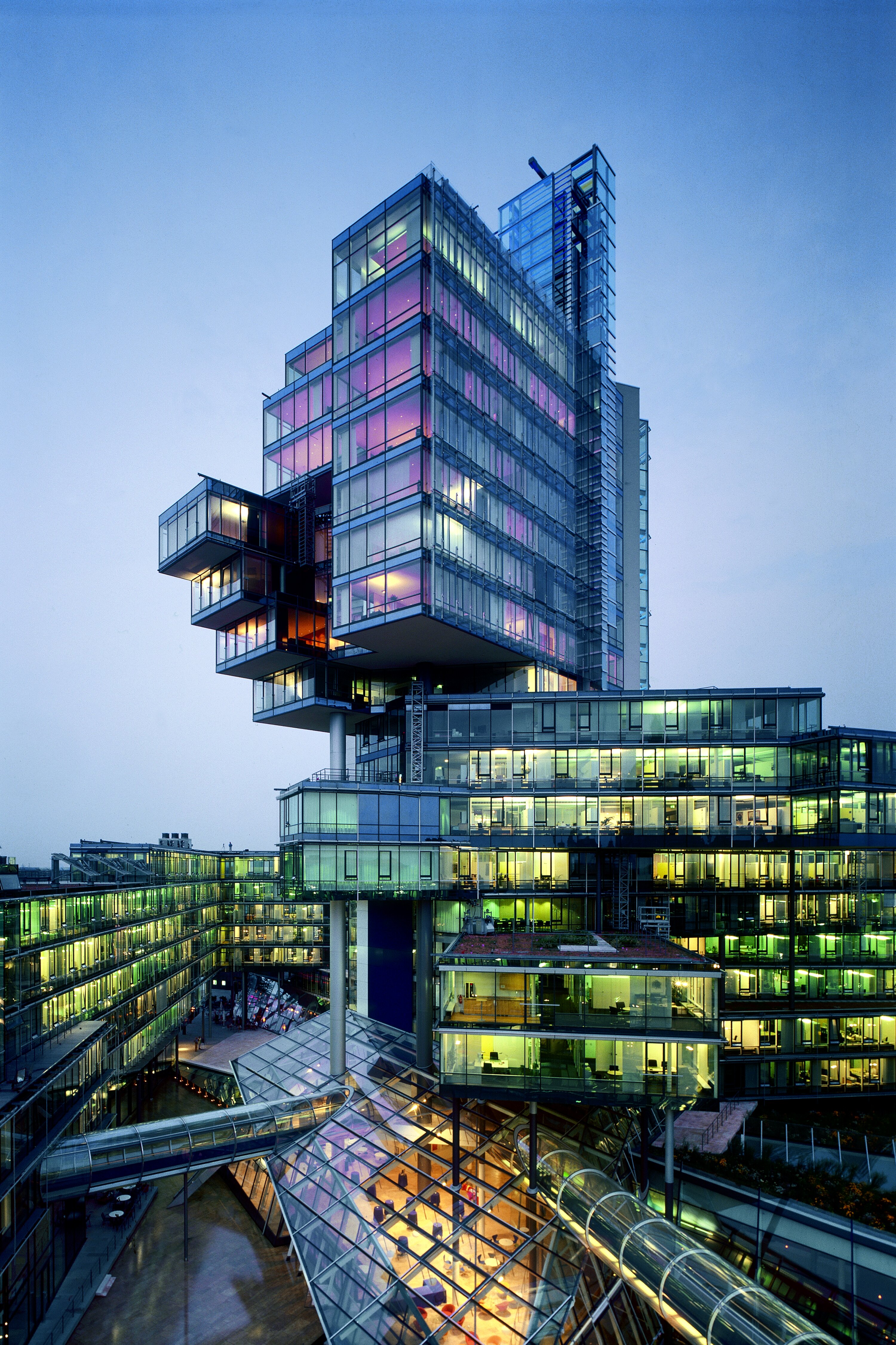 Project by Behnisch Architekturbuero, Norddeutsche Landesbank am Friedrichswall. Illuminated modern glass office tower with stacked volumes at dusk, connected to lower buildings and a glass-roofed atrium below.