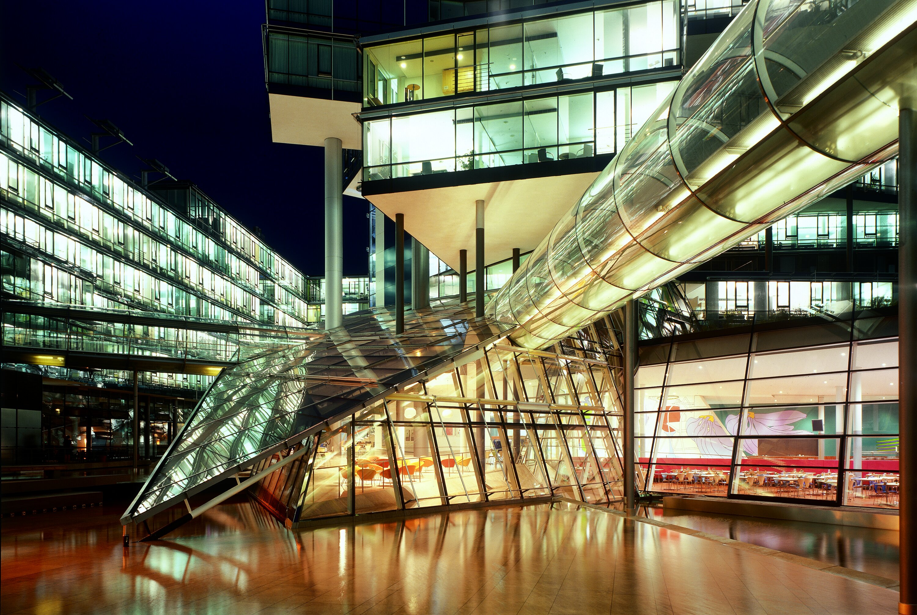 Project by Behnisch Architekturbuero, Norddeutsche Landesbank am Friedrichswall. Night view of the modern glass office complex with illuminated interiors and a tubular skybridge connecting buildings above a lit atrium.