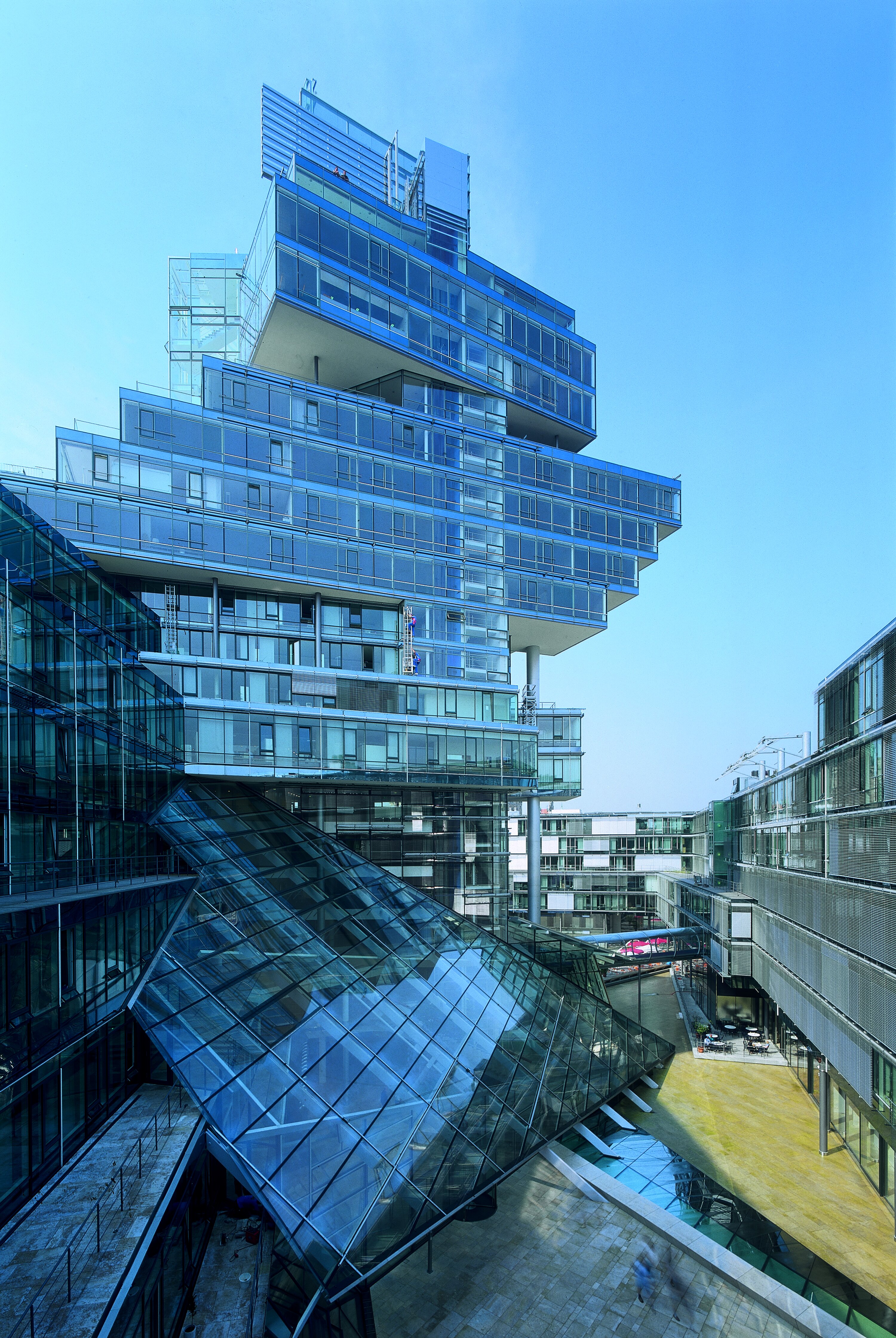Project by Behnisch Architekturbuero, Norddeutsche Landesbank am Friedrichswall. Modern glass office complex with a stacked tower and angular glass atrium roof, surrounded by adjacent buildings under a clear blue sky.