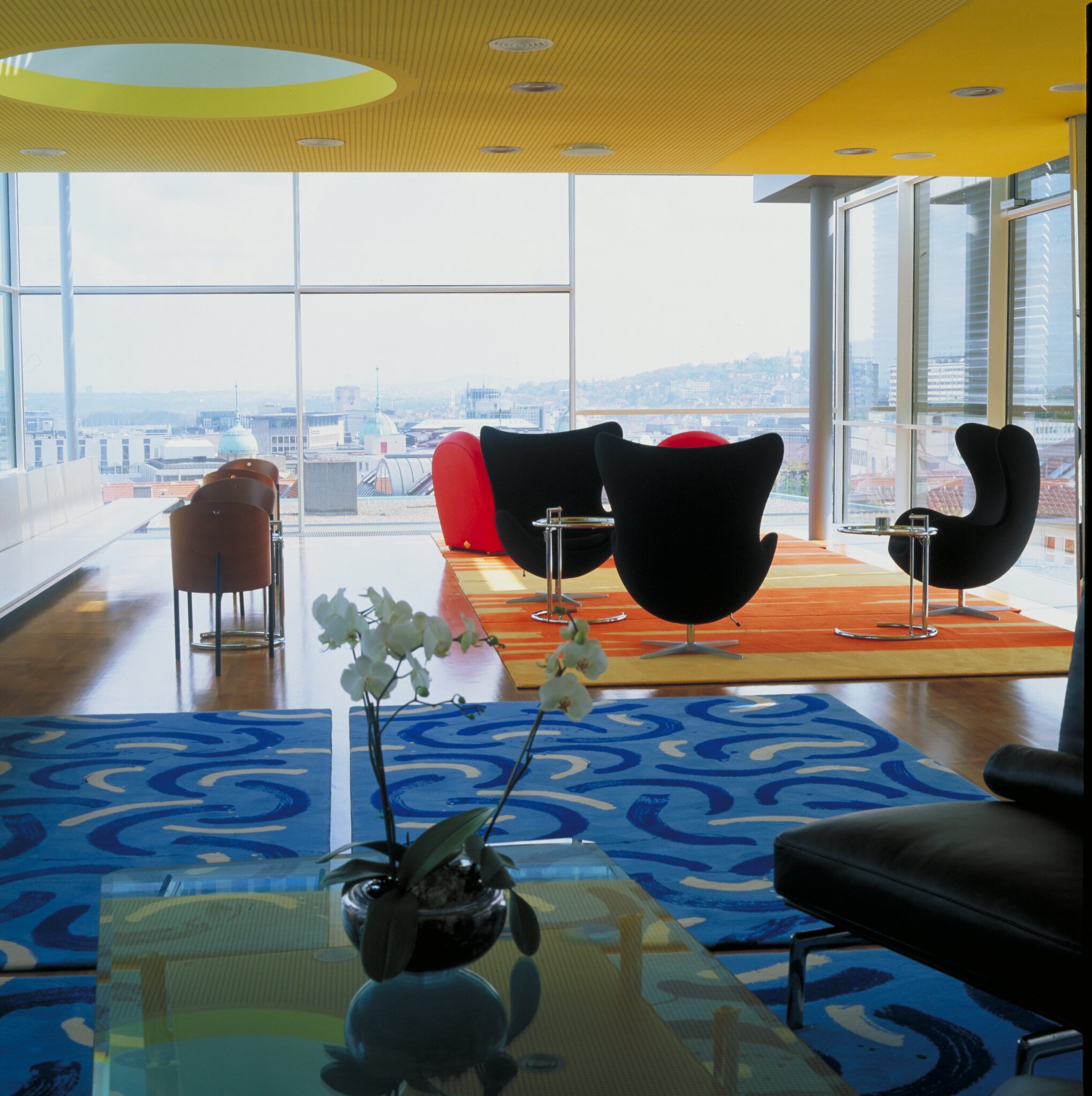 Project by Behnisch Architekturbuero, Landesgirokasse am Bollwerk. Modern lounge with colorful rugs, armchairs, and floor-to-ceiling windows overlooking a city skyline.