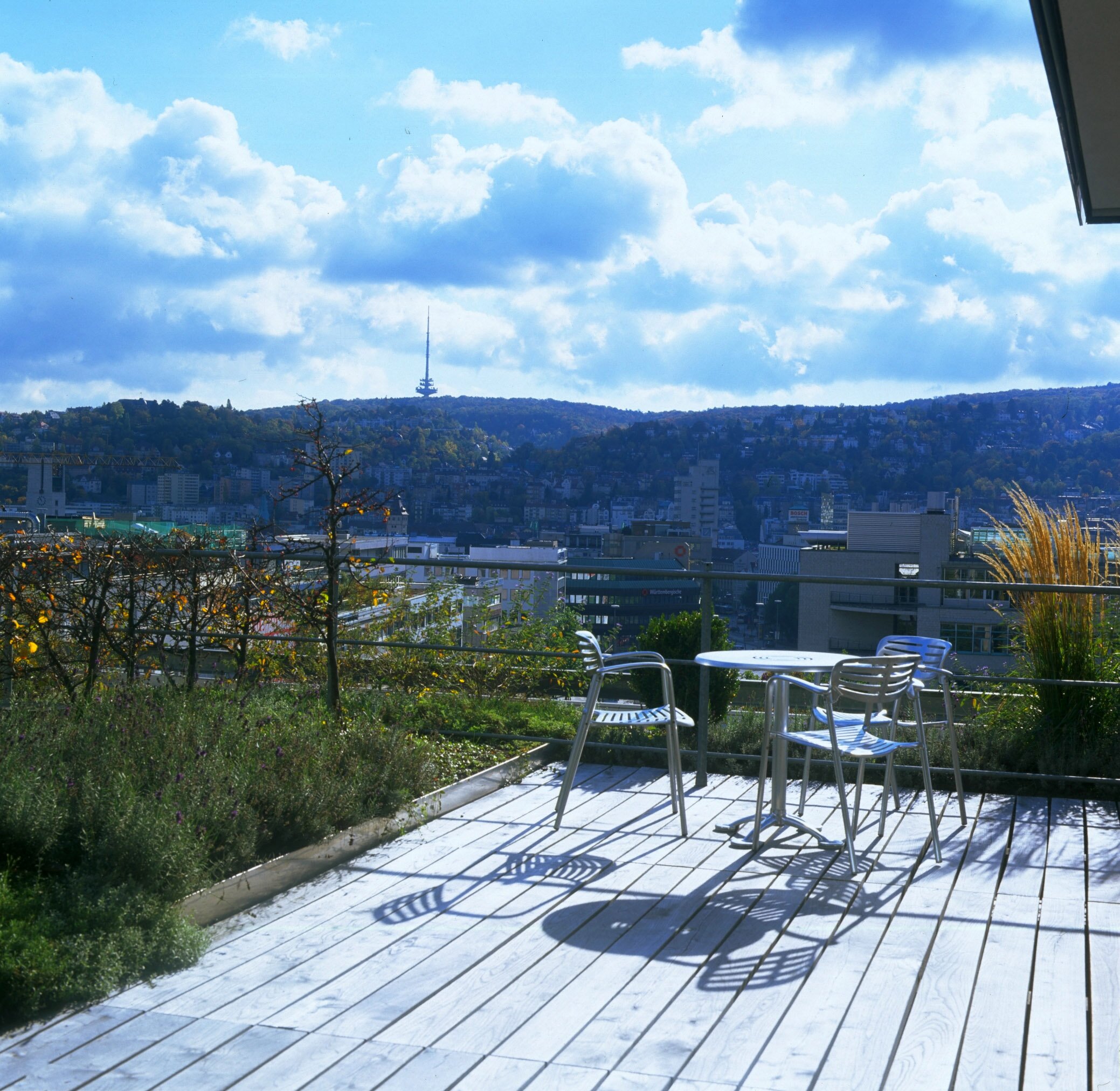 Project by Behnisch Architekturbuero, Landesgirokasse am Bollwerk. Rooftop terrace with metal table and chairs overlooking the city skyline, hills, and a tower under a partly cloudy sky.