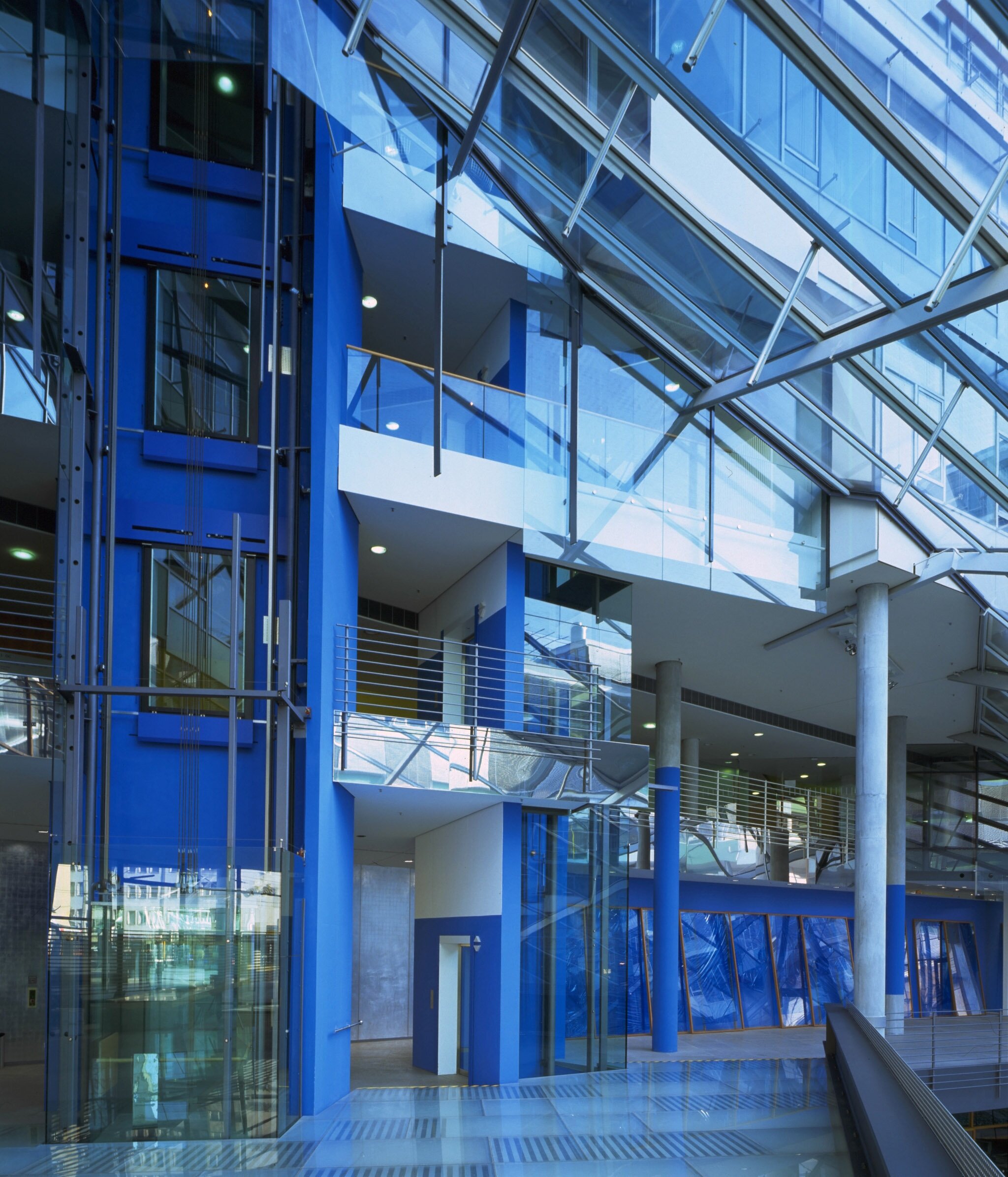 Project by Behnisch Architekturbuero, Landesgirokasse am Bollwerk. Interior of a modern building with blue walls, glass elevator, balconies, and a transparent roof structure letting in daylight.