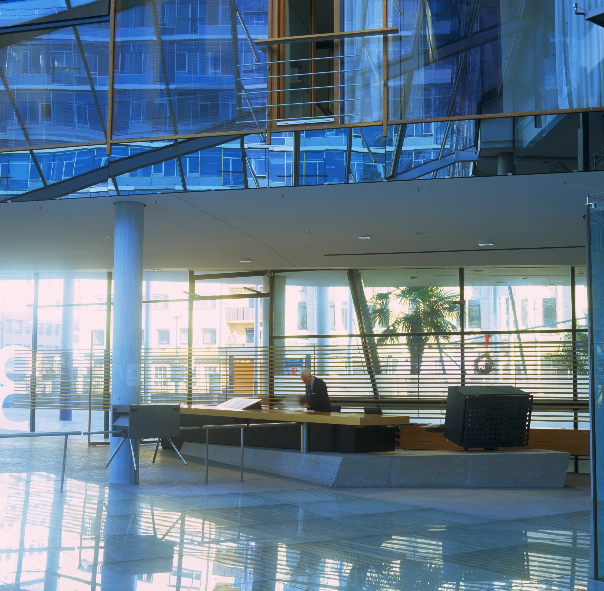 Project by Behnisch Architekturbuero, Landesgirokasse am Bollwerk. Spacious modern lobby with glass walls, reception desk, and a person standing behind the counter, with reflections on the polished floor.