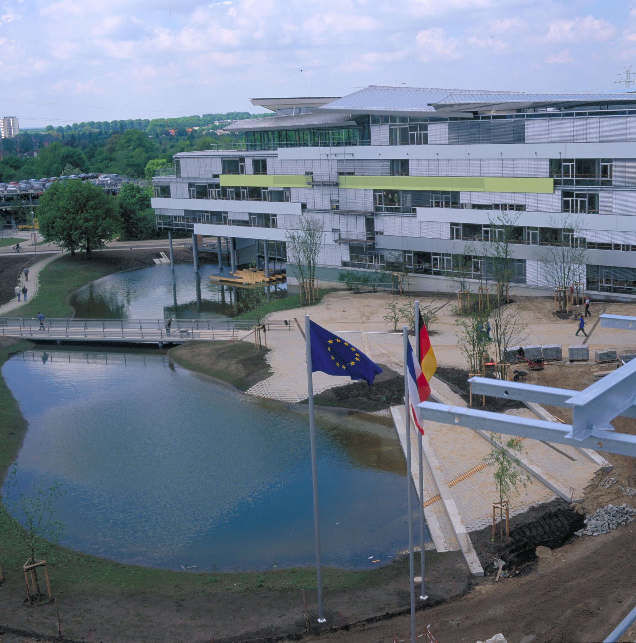 Project by Behnisch Architekturbuero, LVA &ndash; Landesversicherungsanstalt Schleswig-Holstein. Modern office building beside a pond with footbridge, landscaped paths, and EU and German flags in the foreground.
