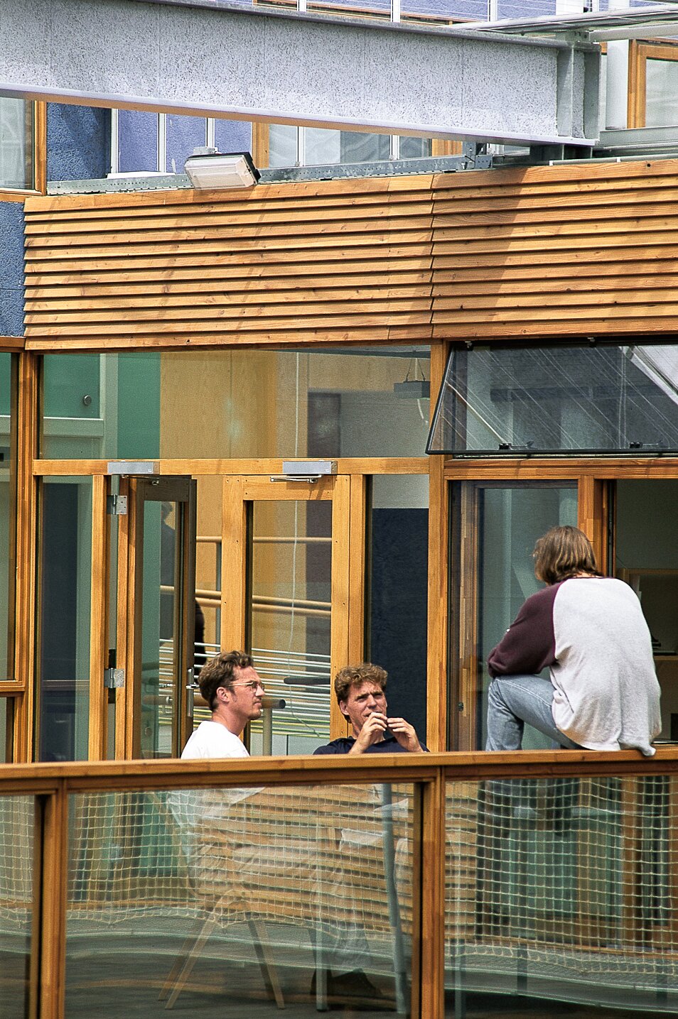 Project by Behnisch Architekturbuero, IBN-Institute for Forestry and Nature Research. Three people are talking on a wooden balcony in front of a building with glass doors and windows; one person is sitting on the railing, the others on chairs.