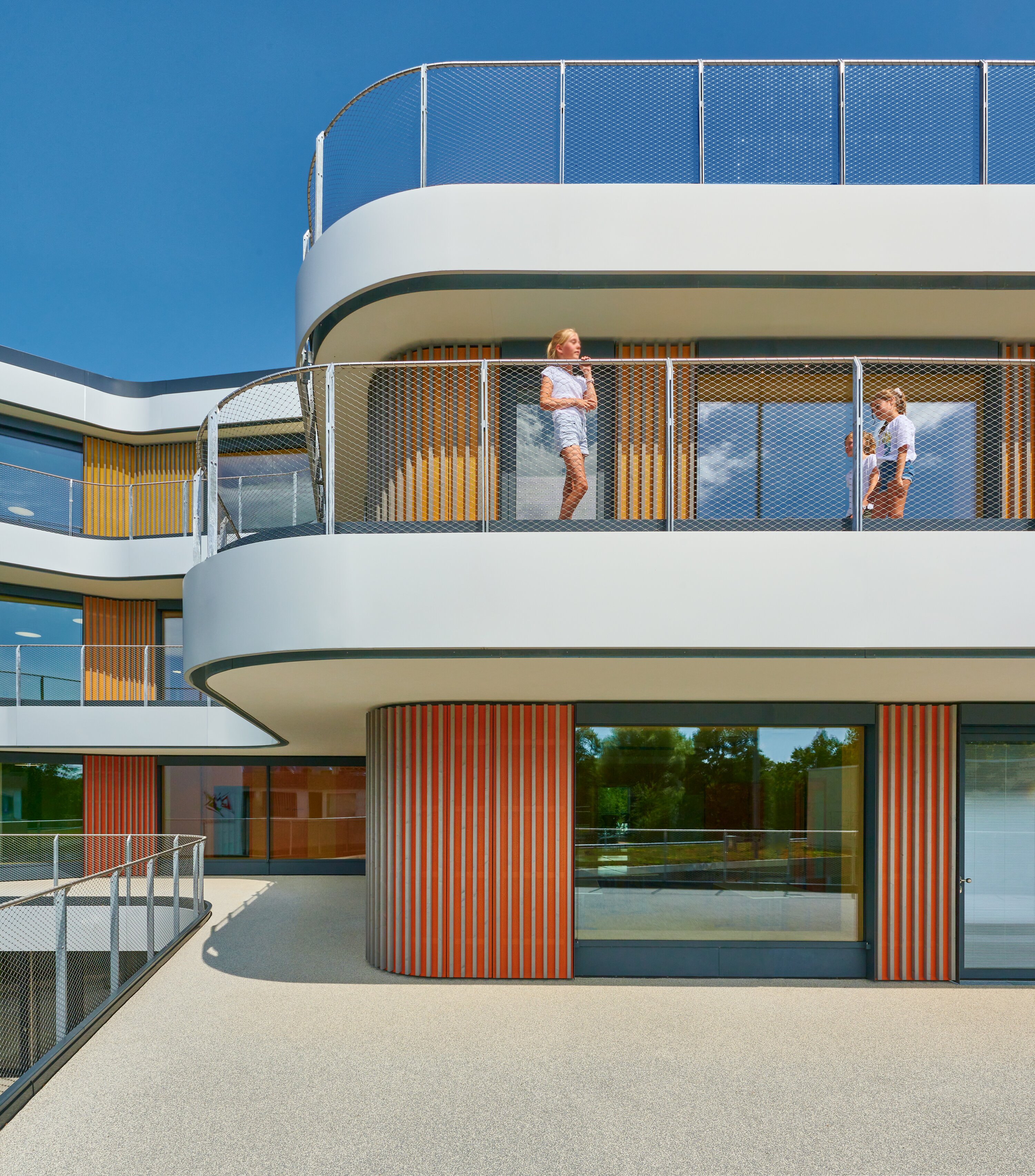 Project by Behnisch Architekturbuero, Gotthard-M&uuml;ller-School Bernhausen. Two children stand on a curved balcony of the building with glass railing and colorful vertical facade elements.