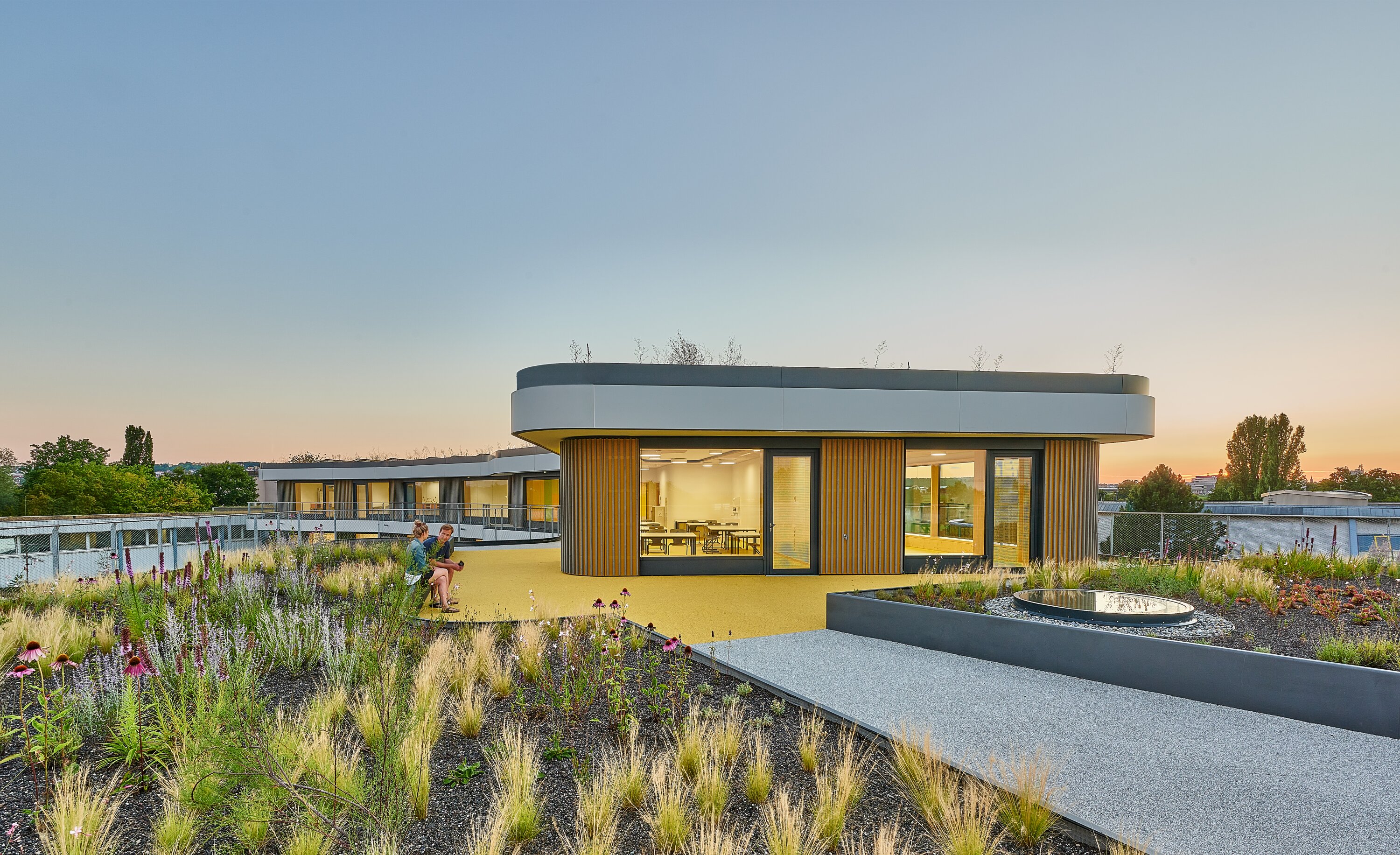 Project by Behnisch Architekturbuero, Gotthard-M&uuml;ller-School Bernhausen. A rooftop terrace with a landscaped garden and pathways; two people sit next to a modern, circular building with large windows at sunset. Part of the flooring is marked in yellow.