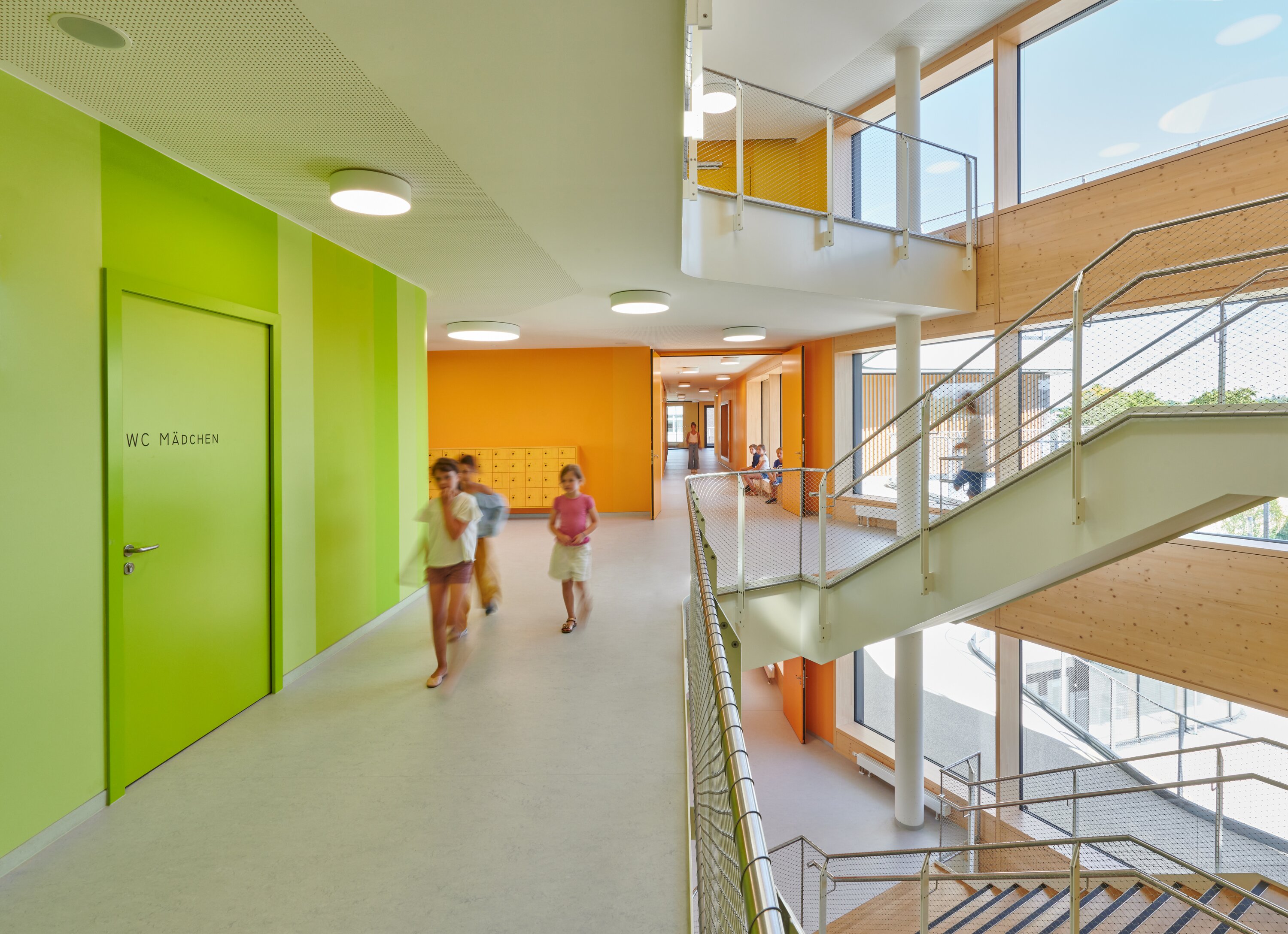 Project by Behnisch Architekturbuero, Gotthard-M&uuml;ller-School Bernhausen. Bright school hallway with green and orange walls, children walking, and an open stairwell with metal railings and large windows.