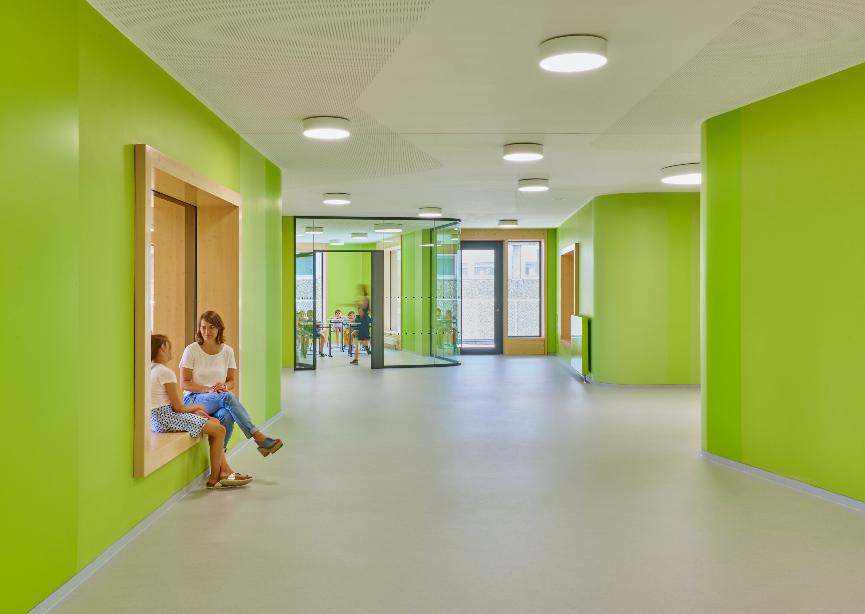 Project by Behnisch Architekturbuero, Gotthard-M&uuml;ller-School Bernhausen. Bright green school hallway with curved walls, a teacher and a student sitting in a window niche, and a classroom visible through glass doors.