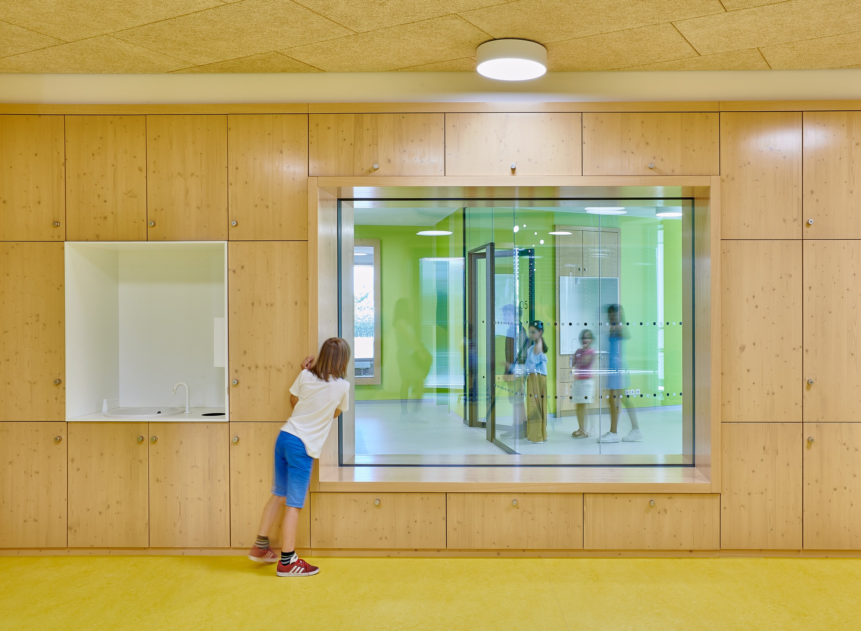 Project by Behnisch Architekturbuero, Gotthard-M&uuml;ller-School Bernhausen. Child leans on wood-paneled wall, looking through large interior window into classroom with children in bright green room.
