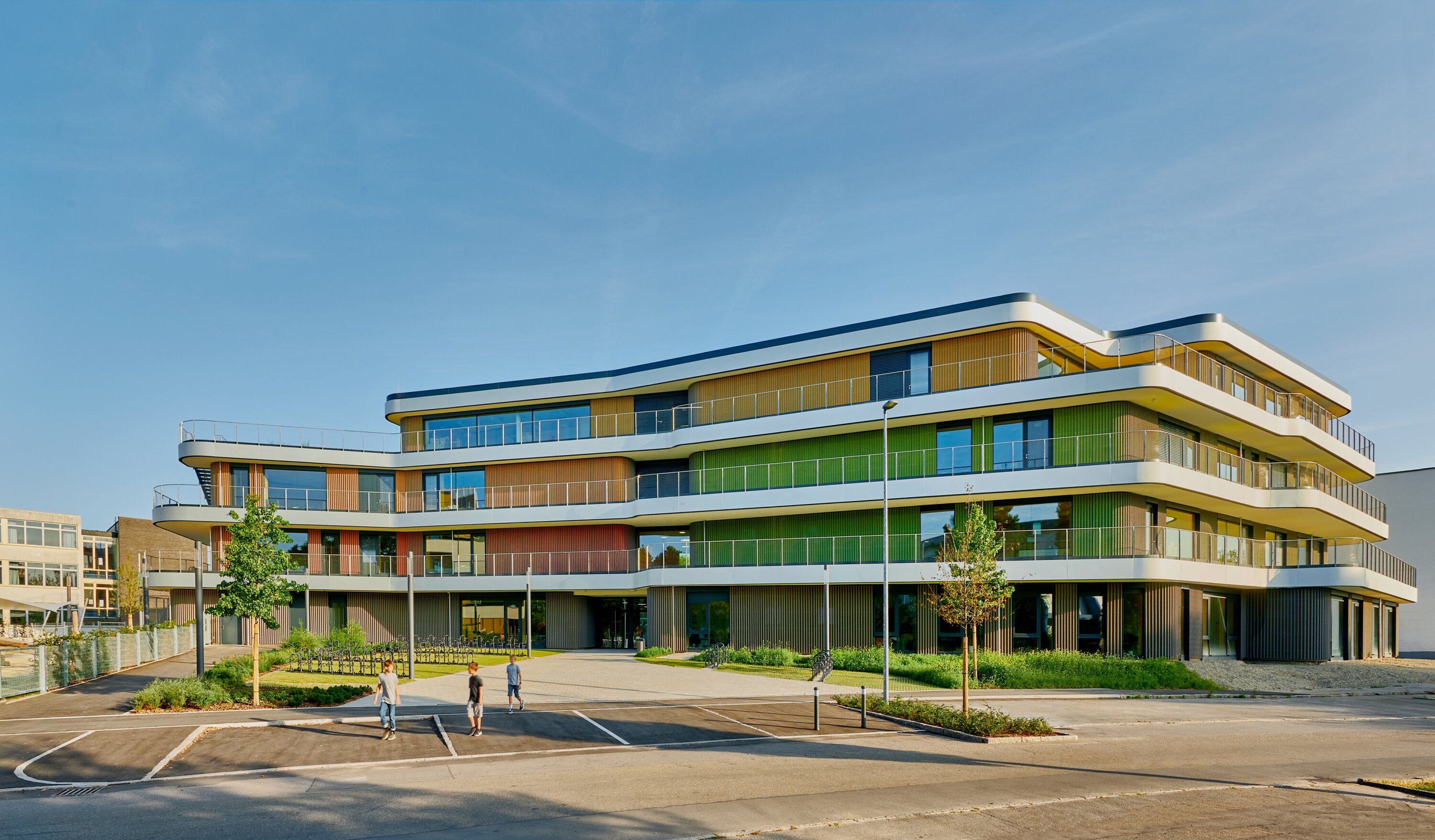 Project by Behnisch Architekturbuero, Gotthard-M&uuml;ller-School Bernhausen. Multi-story schoolbuilding with curved balconies and colorful facade, set by a street with a few people walking nearby.