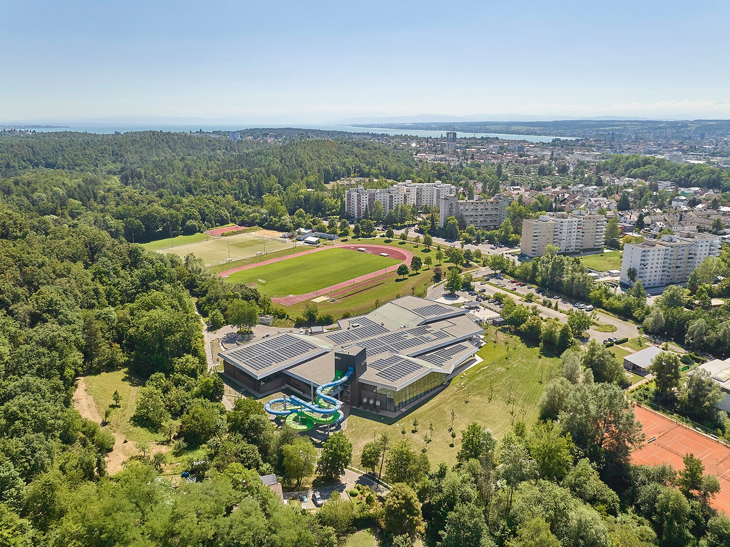 Project by Behnisch Architekturbuero, Schwaketenbad. Aerial view of the building with water slides. A photovoltaic system is located on the roof. Nearby are a running track, residential buildings, a forest, and a lake in the distance.