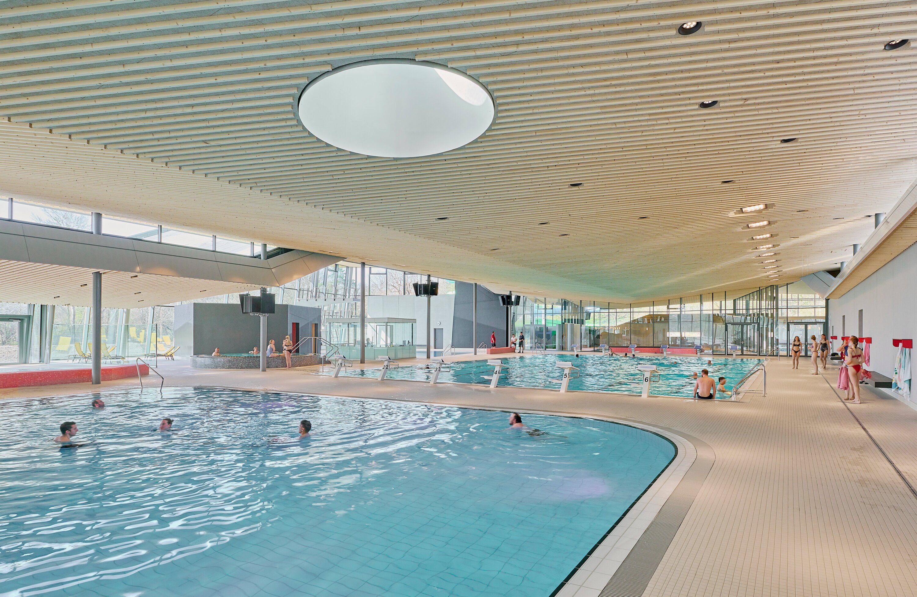 Project by Behnisch Architekturbuero, Schwaketenbad. An indoor swimming pool with several pools, where people swim and stand around, beneath a large wooden slatted ceiling. The swimming hall is flooded with light through the large glass facade and feels open.
