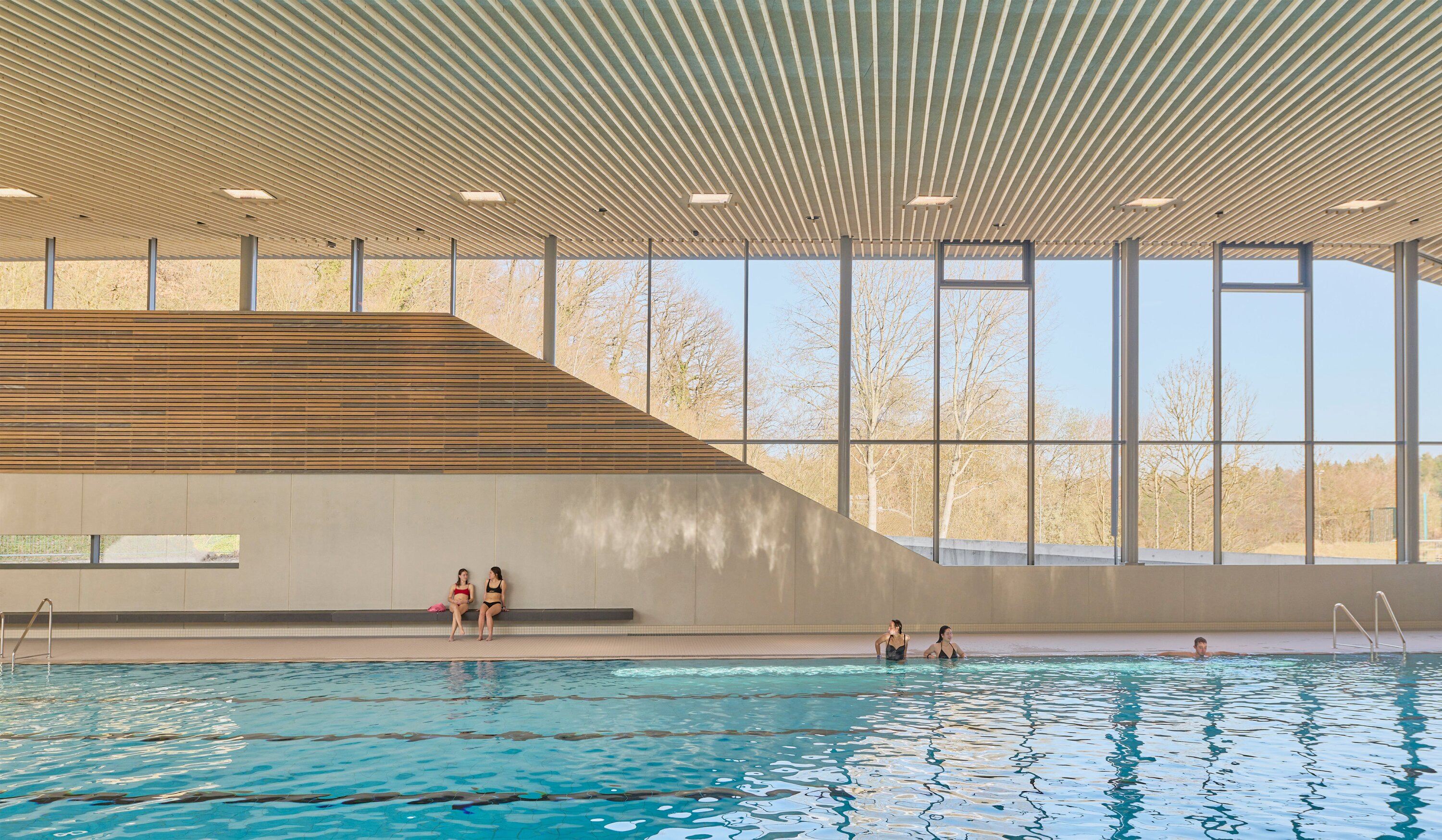 Project by Behnisch Architekturbuero, Sportbad Friedrichshafen. Courtyard with glass buildings around a pool, landscaped garden with trees and umbrellas, and terraces under a partly cloudy sky.