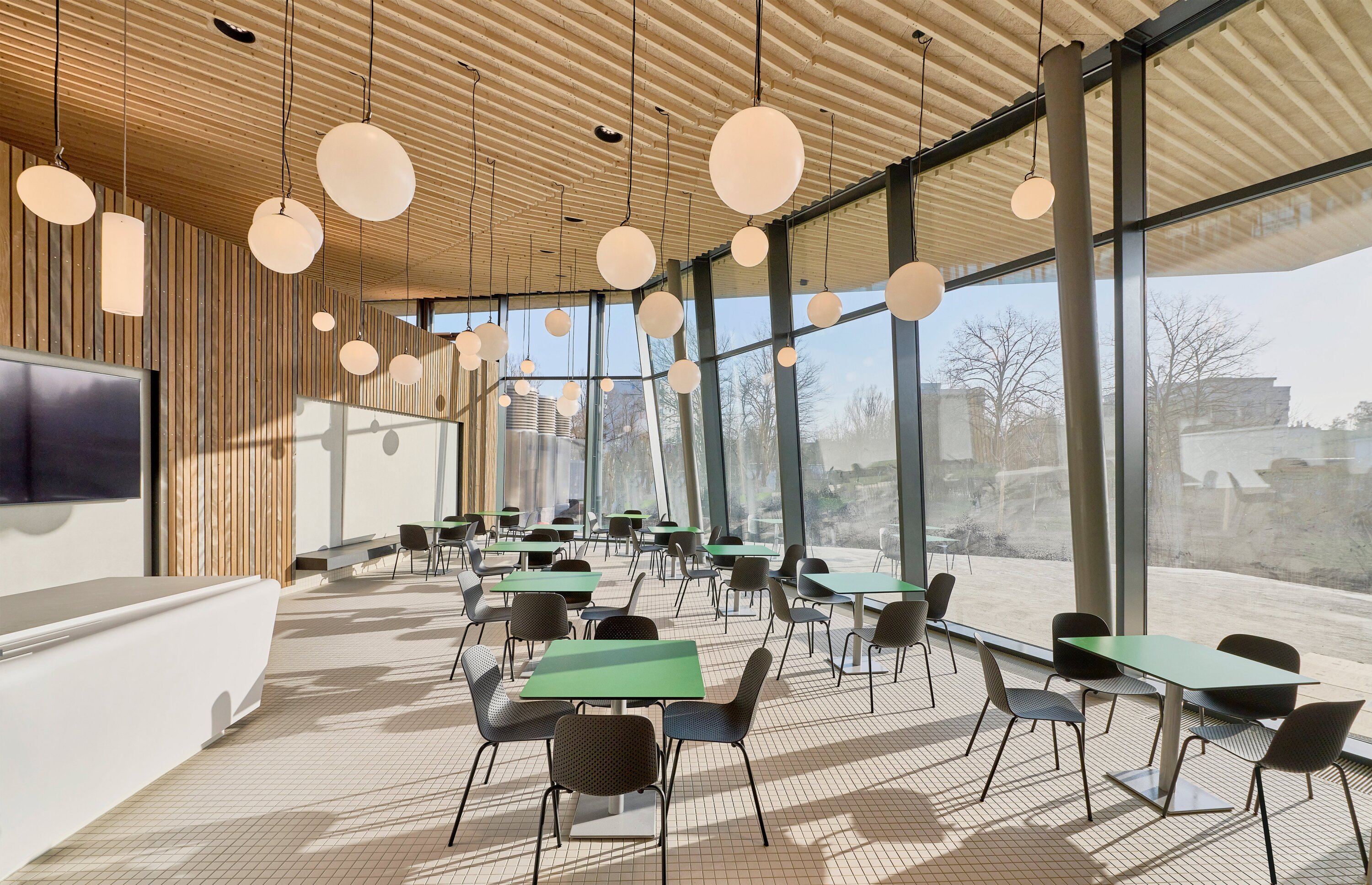 Project by Behnisch Architekturbuero, Schwaketenbad. Bright caf&eacute; interior with green tables and black chairs, large glass walls, wood ceiling, and hanging globe lights.