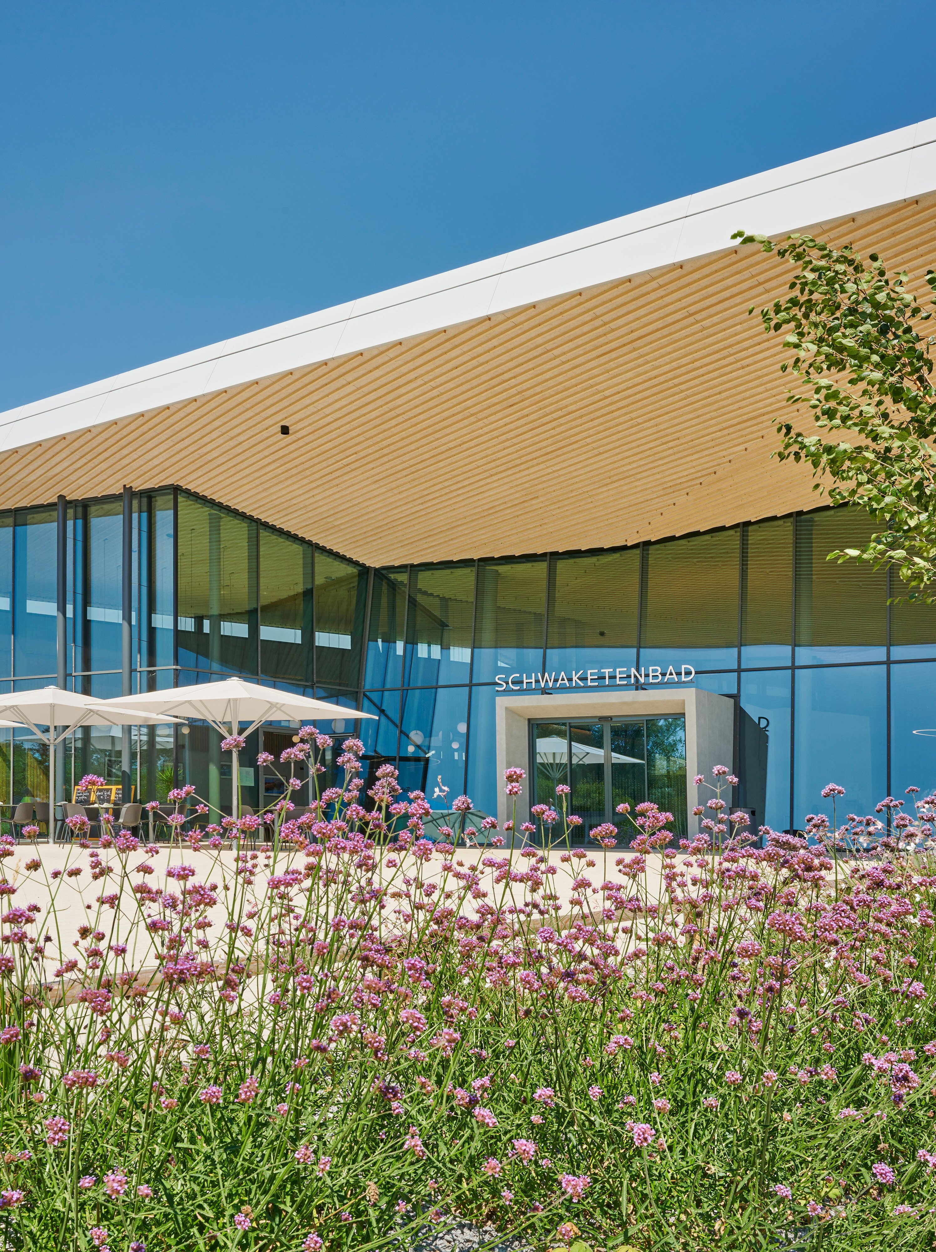 Project by Behnisch Architekturbuero, Schwaketenbad. Glass-fronted building entrance labeled &ldquo;Schwaketembad,&rdquo; with wooden overhang and flowers in the foreground.