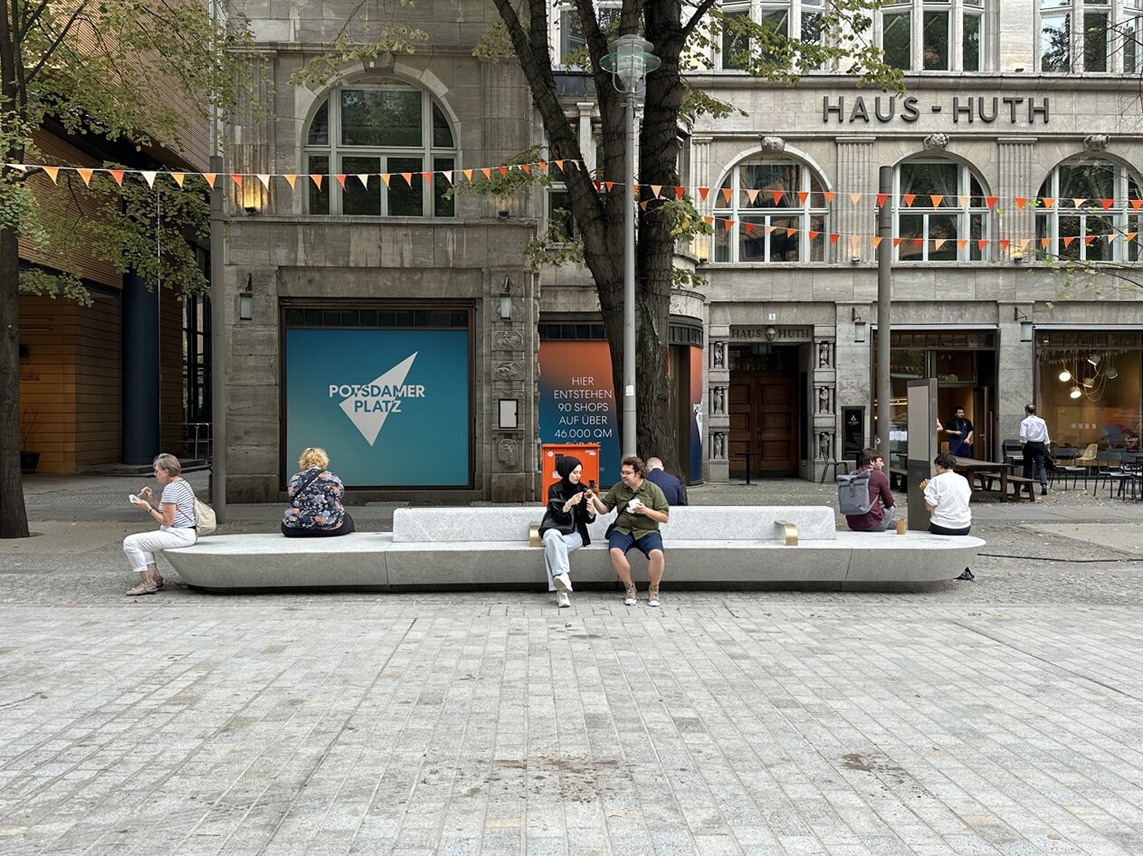 Project by Behnisch Architekturbuero, Potsdamer Platz Quartier. People are sitting on a spacious bench next to a pedestrian zone; shops are visible in the background.