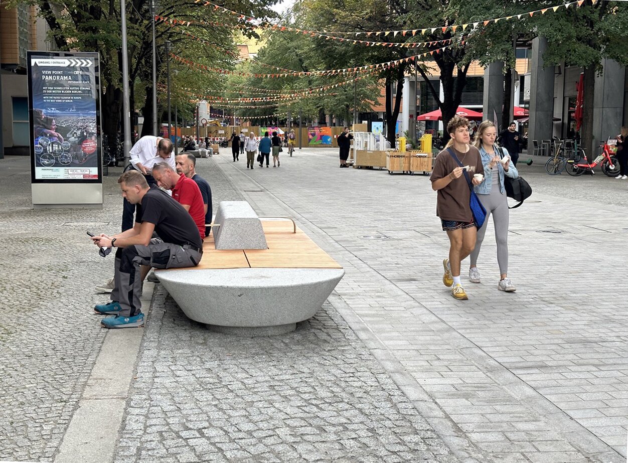 Project by Behnisch Architekturbuero, Potsdamer Platz Quartier. Three men are sitting on a spacious bench next to a pedestrian zone and take a break. A couple is walking near by on the right hand side.