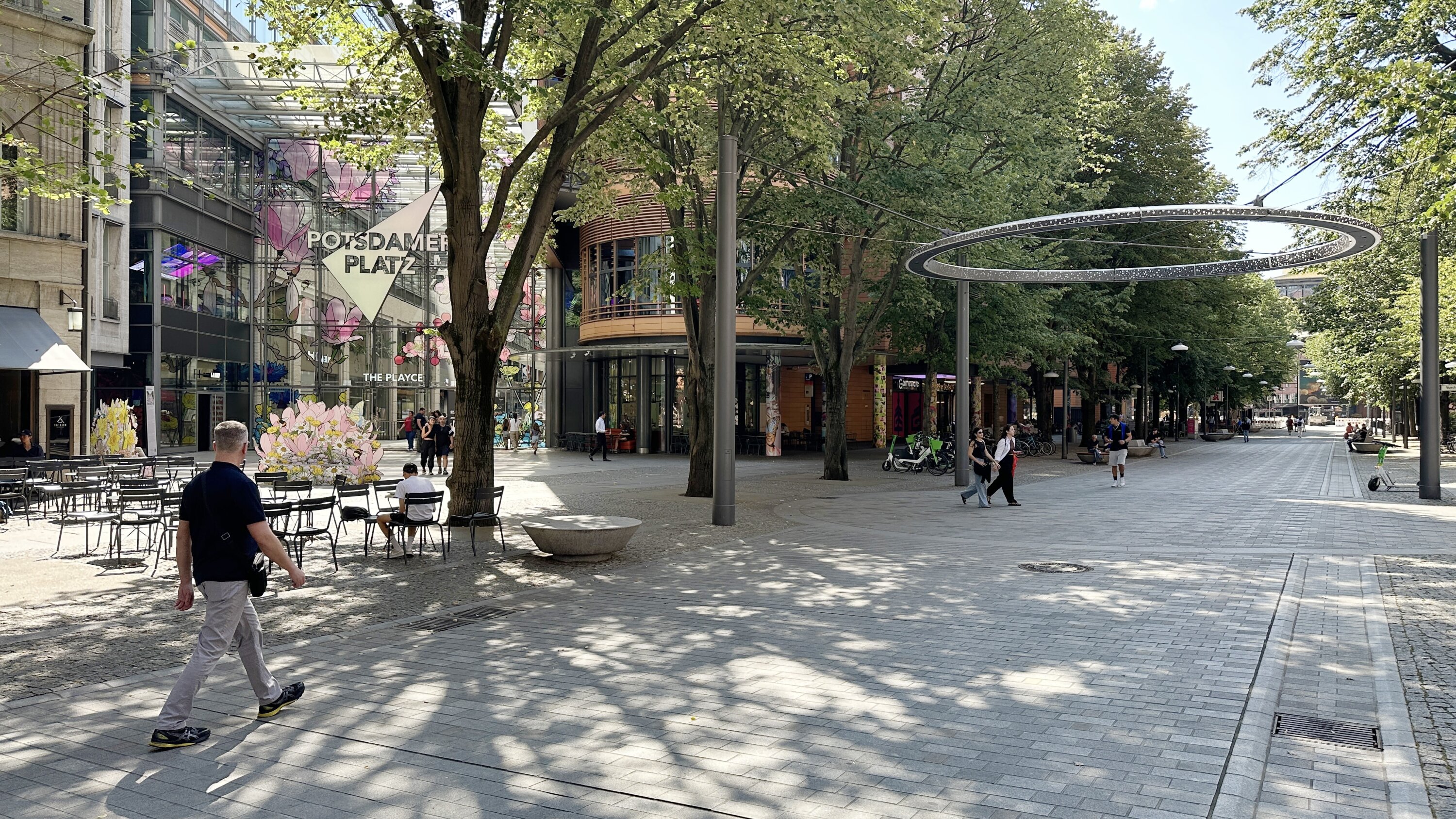 Project by Behnisch Architekturbuero, Potsdamer Platz Quartier. The tree-lined pedestrian zone at Potsdamer Platz with cafes, seating areas and pedestrians. The glass entrance can be seen in the background.