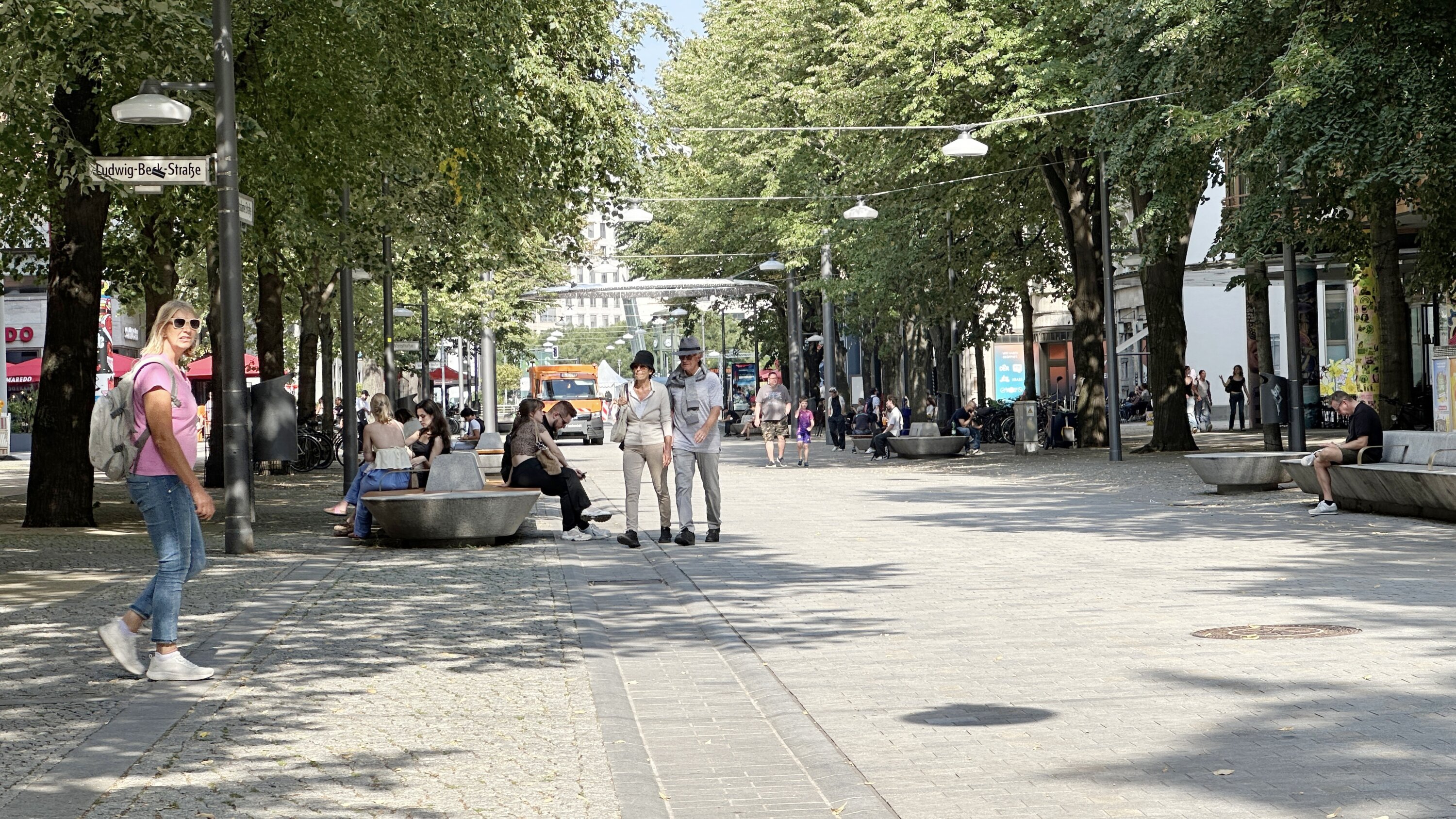 Project by Behnisch Architekturbuero, Potsdamer Platz Quartier. A pedestrian zone lined with trees, with people strolling and shops on both sides. People are sitting on the benches next to the pedestrian zone.
