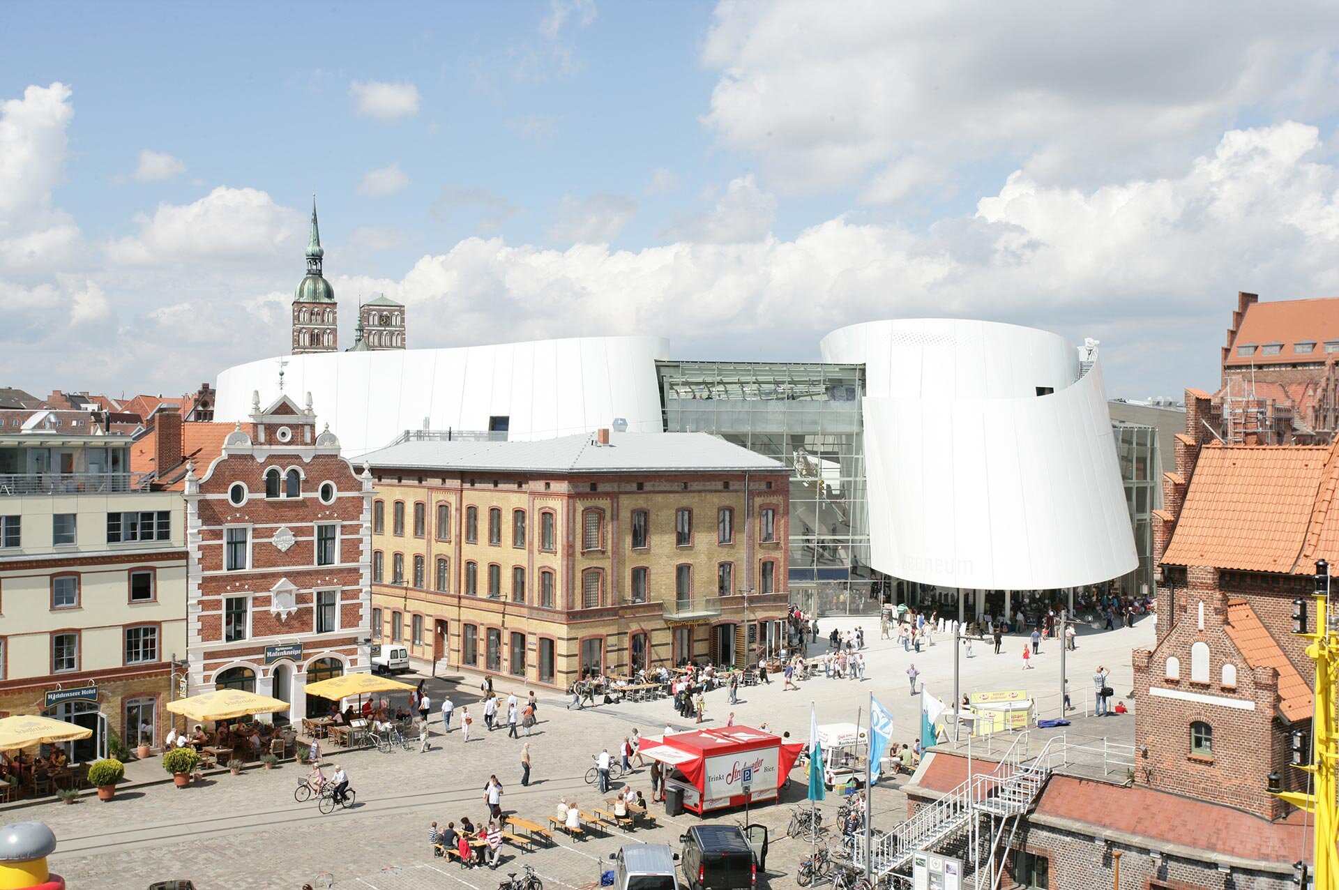 Project by Behnisch Architekturbuero, Ozeanum. City square with historic brick buildings and a modern white museum, with people walking, cycling, and gathering outdoors.