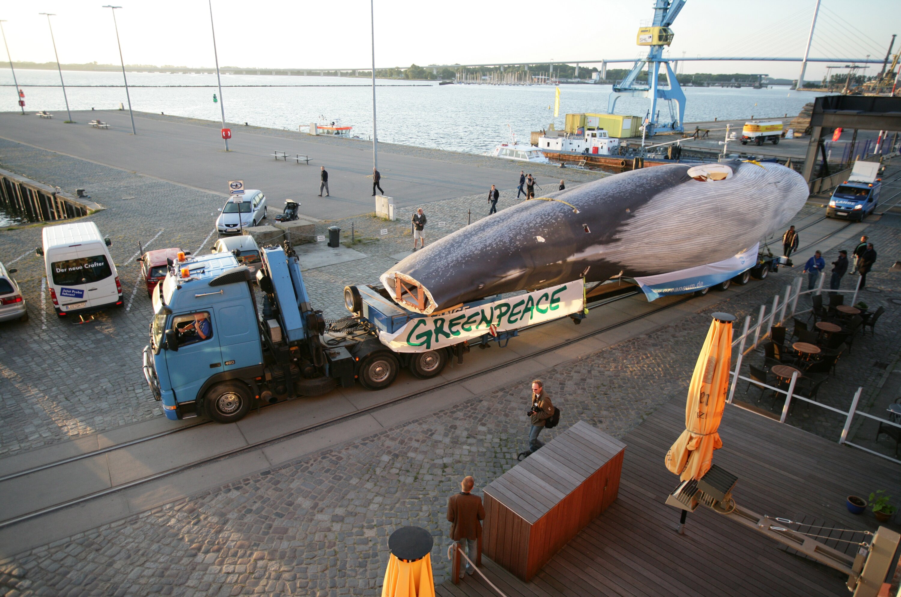 Project by Behnisch Architekturbuero, Ozeanum. Truck transporting a large whale model  along a waterfront, with people, cranes, and harbor in the background.