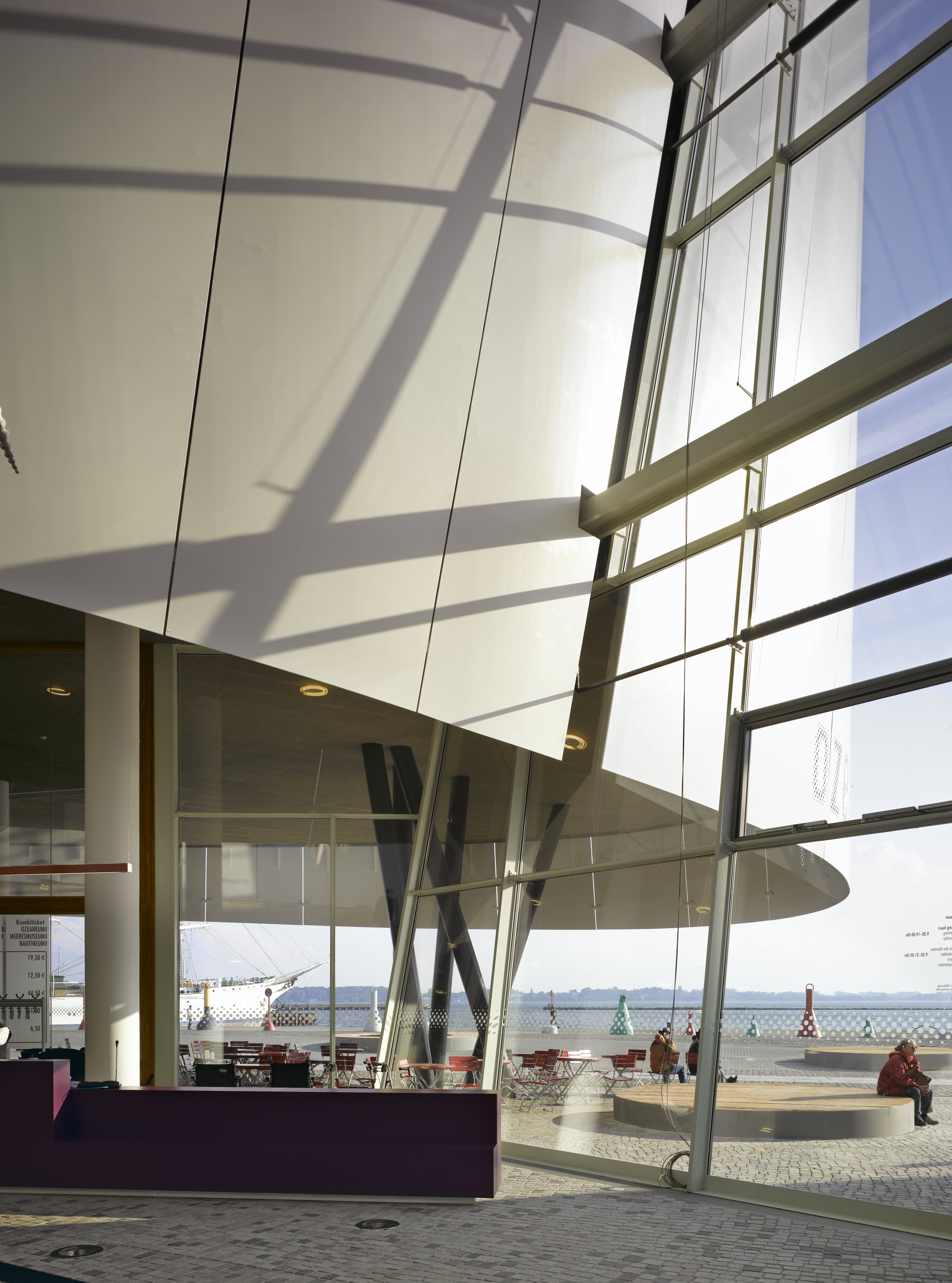 Project by Behnisch Architekturbuero, Ozeanum. Interior view with a large, sloping glass facade; sunlight casts shadows. The glass facade is interrupted by the rounded white structure, which cantilevers outwards. Outside, a terrace by the water with seating and people can be seen.
