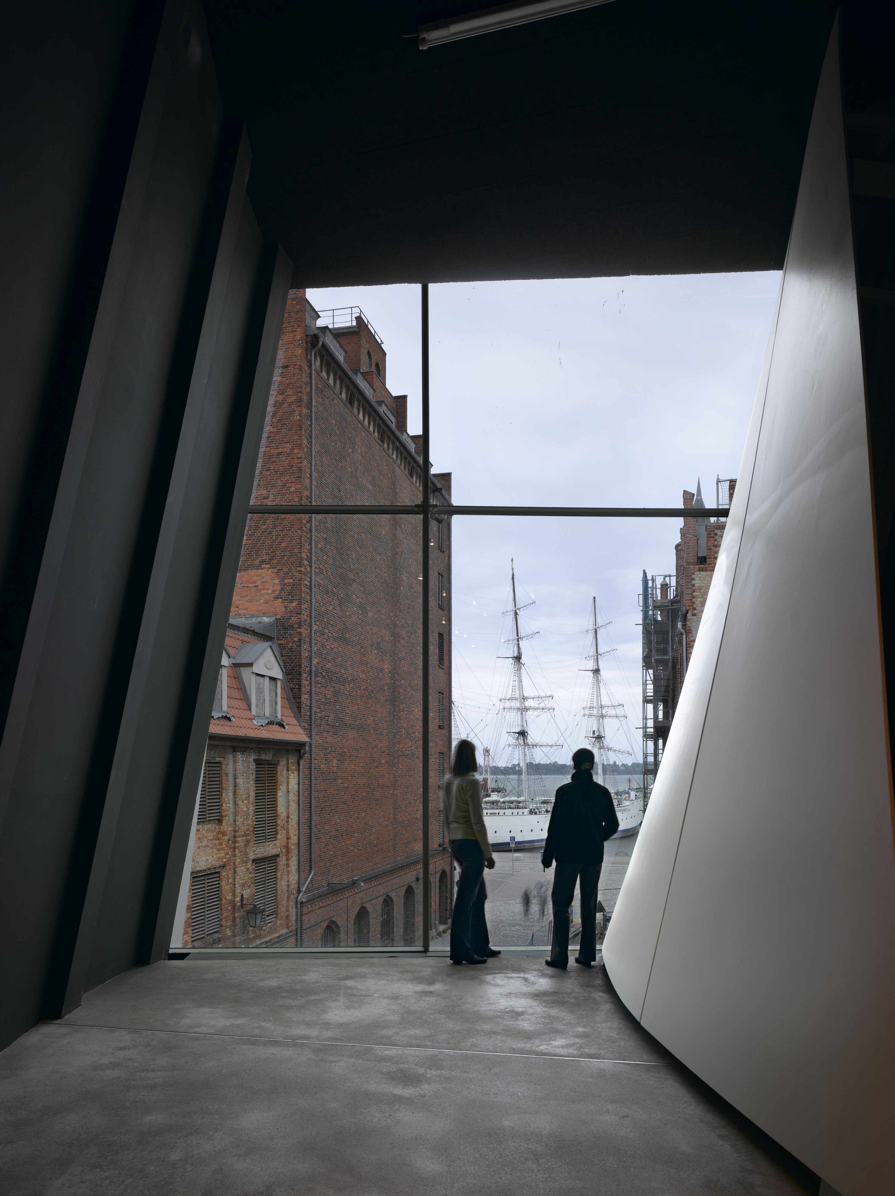 Project by Behnisch Architekturbuero, Ozeanum. Two people stand by a large window, looking out at historic brick buildings and tall ships in the harbor.