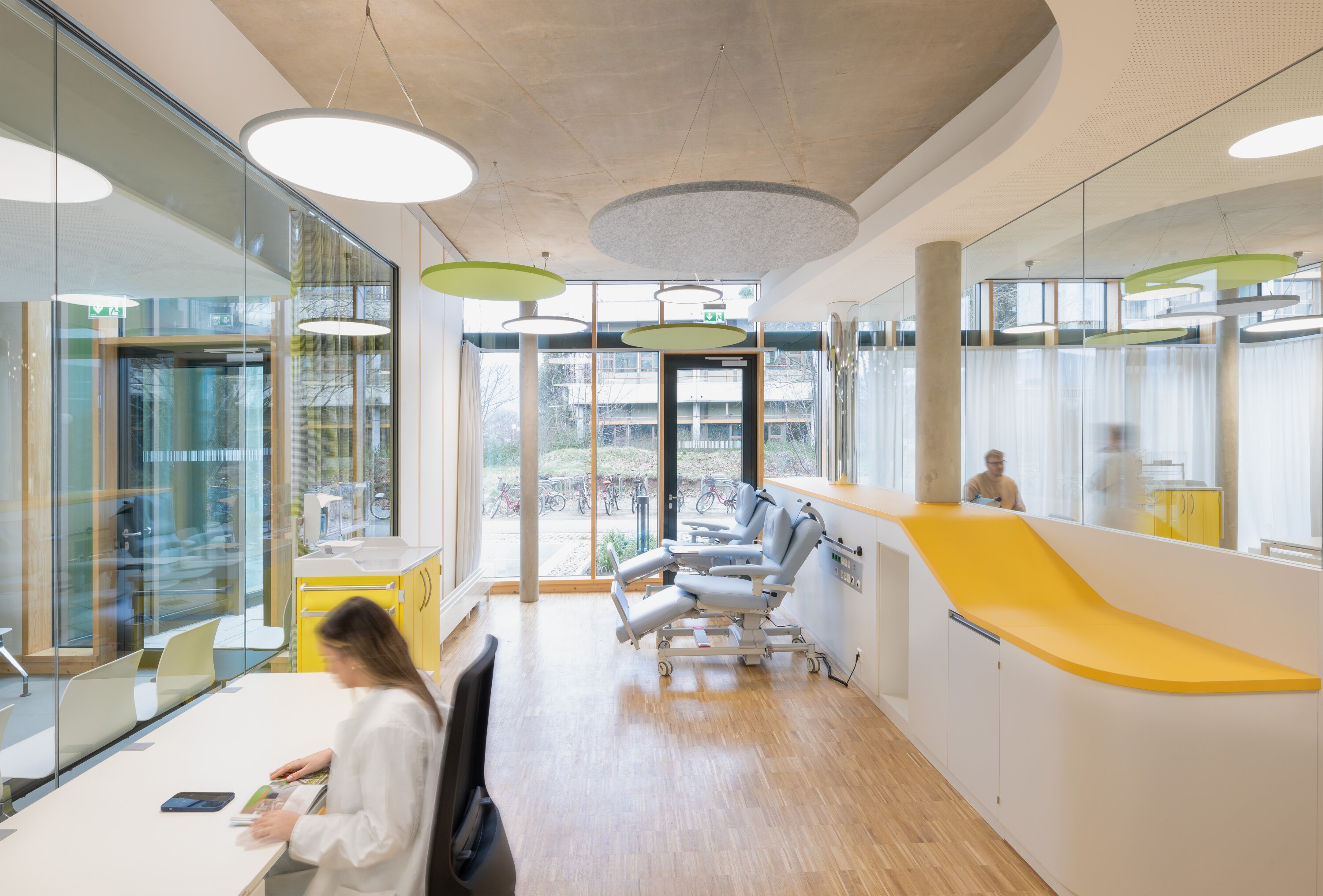 Project by Behnisch Architekturbuero, NCT New Building and Expansion. Treatment room with treatment chairs, glass partitions, and staff at desks in a bright, modern interior for the healthcare sector. Mobile pedestals and work surfaces are colored signal yellow.