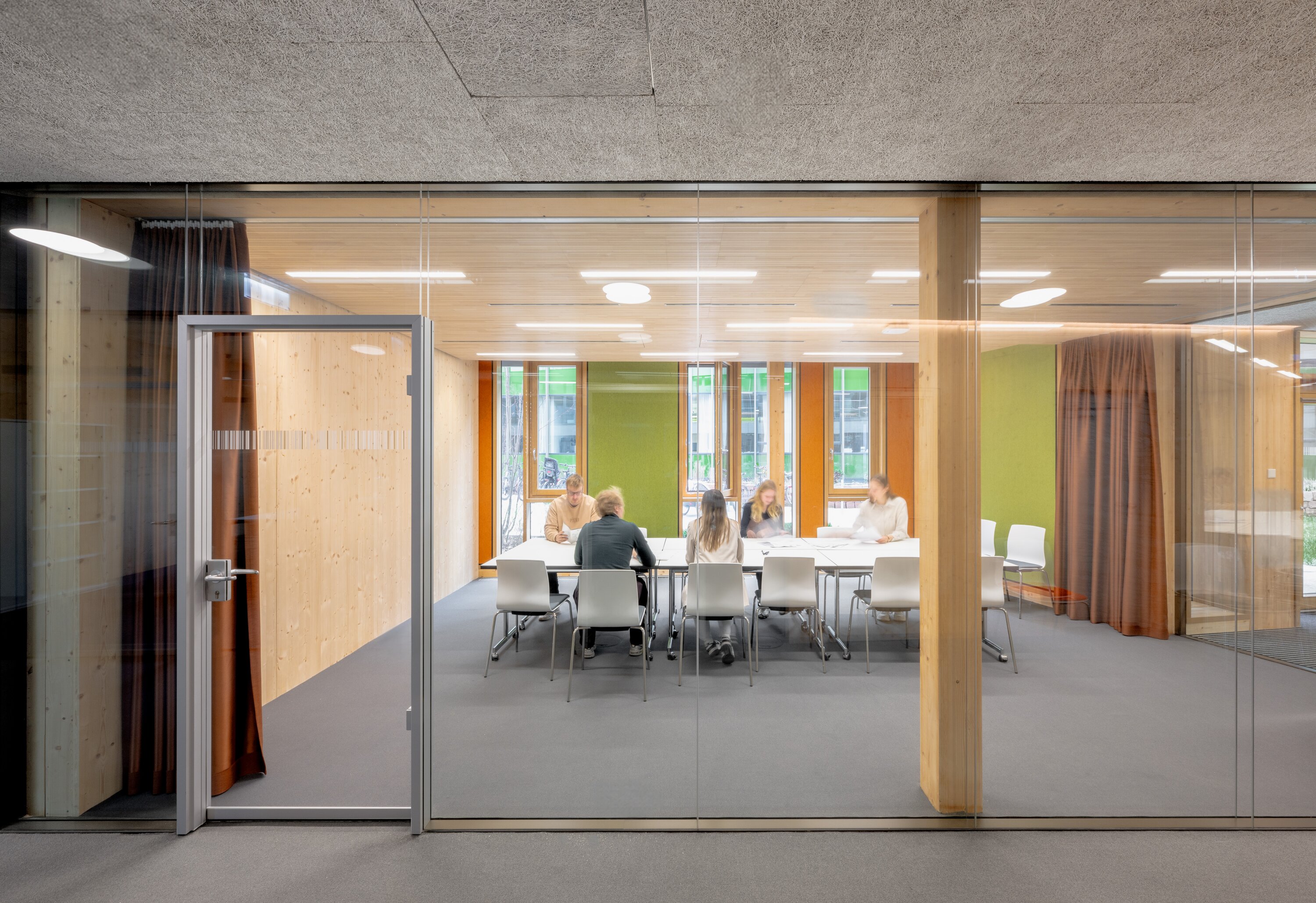 Project by Behnisch Architekturbuero, NCT New Building and Expansion. A meeting room with glass and wood walls, where a group is seated around a table. Colorful accents are provided by orange and green wall panels and orange/ochre curtains.