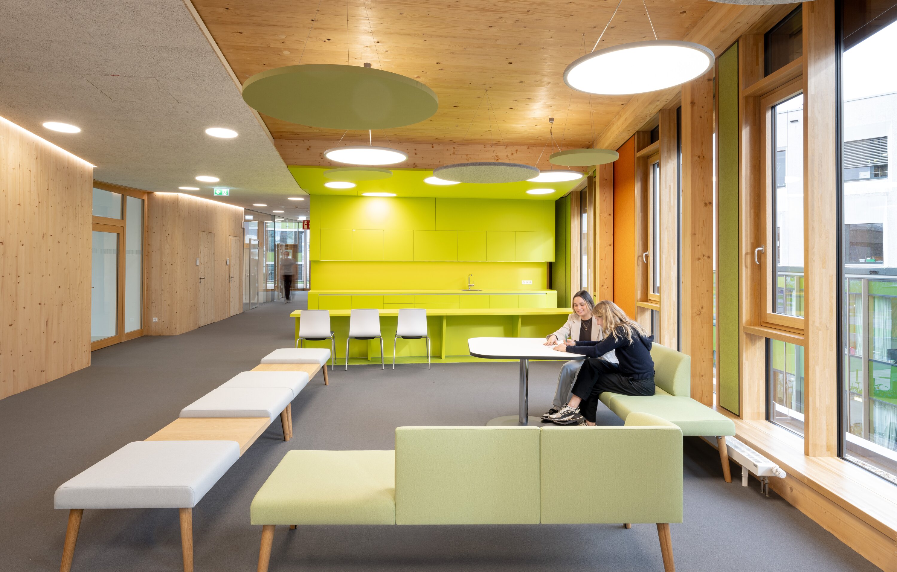 Project by Behnisch Architekturbuero, NCT New Building and Expansion. A bright lounge with a green kitchenette, wooden walls, benches, and two people sitting at a table by large windows. To the left of the picture, a hallway is visible leading to other parts of the building.