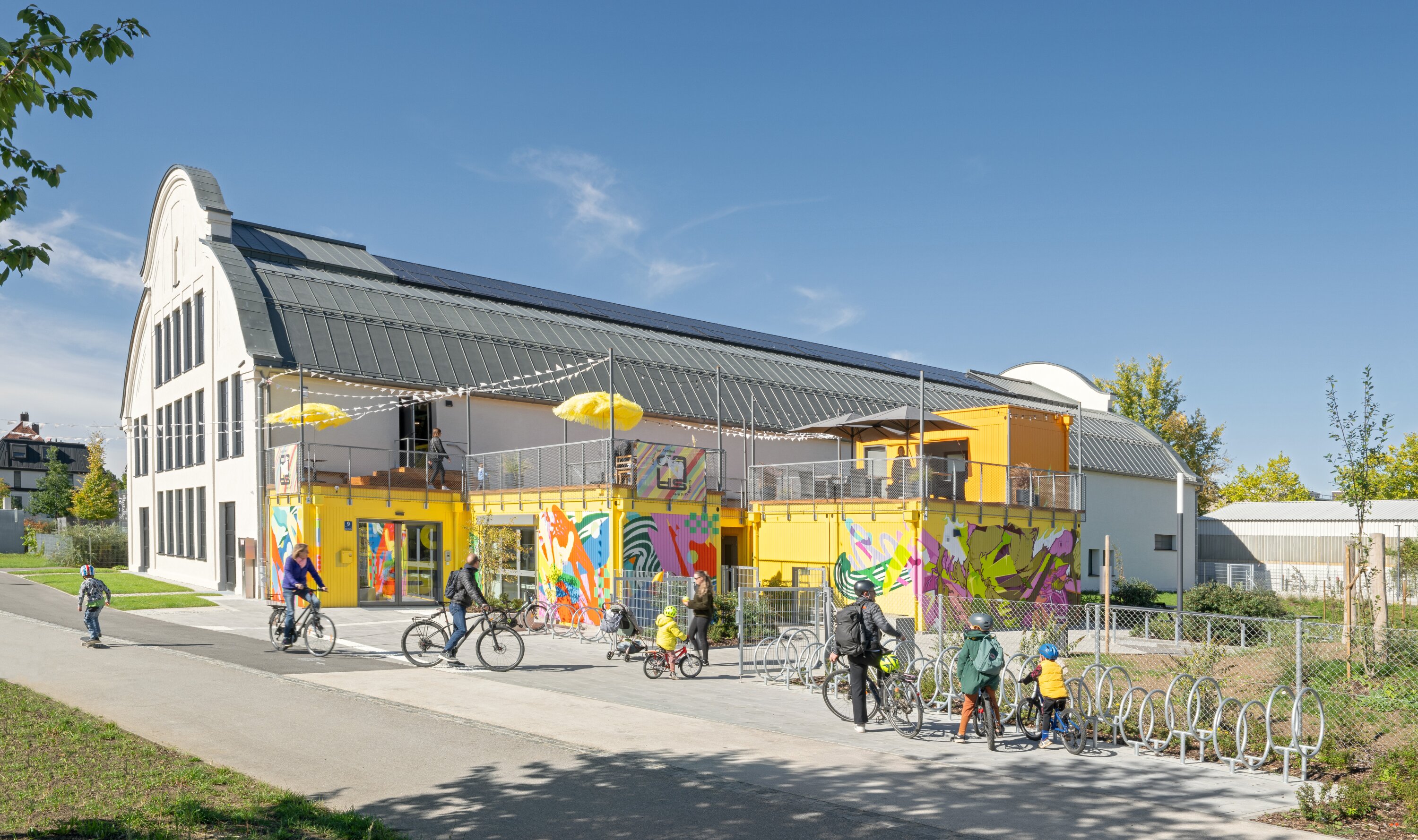 Project by Behnisch Architekturbuero, Action Sport Center. A hall with a container-like extension, a terrace, and people with bicycles in front of it. The containers are yellow and also painted in bright colors.