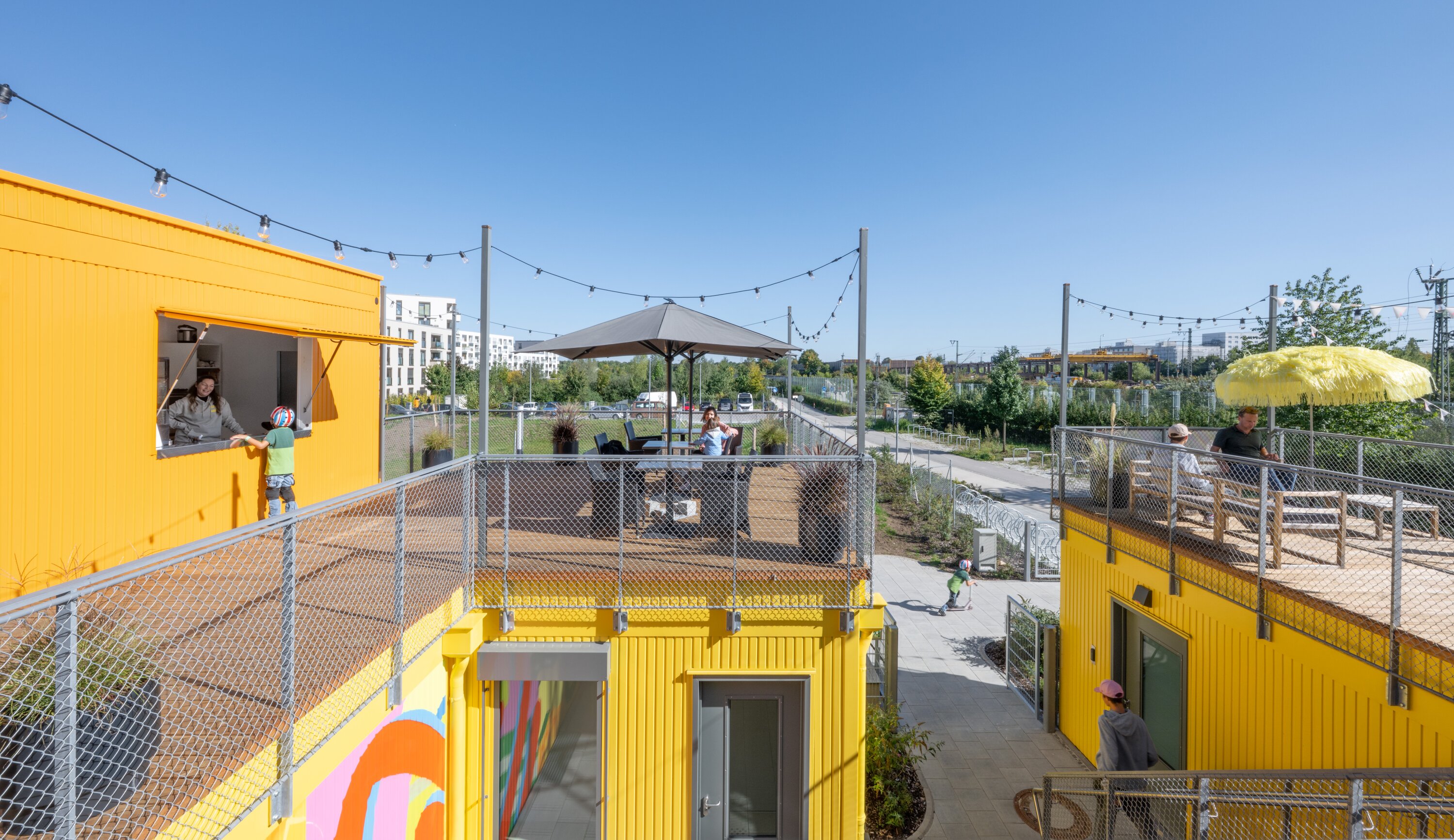 Project by Behnisch Architekturbuero, Action Sport Center. Yellow container-like terraced buildings with seating, people and strings of lights outdoors.