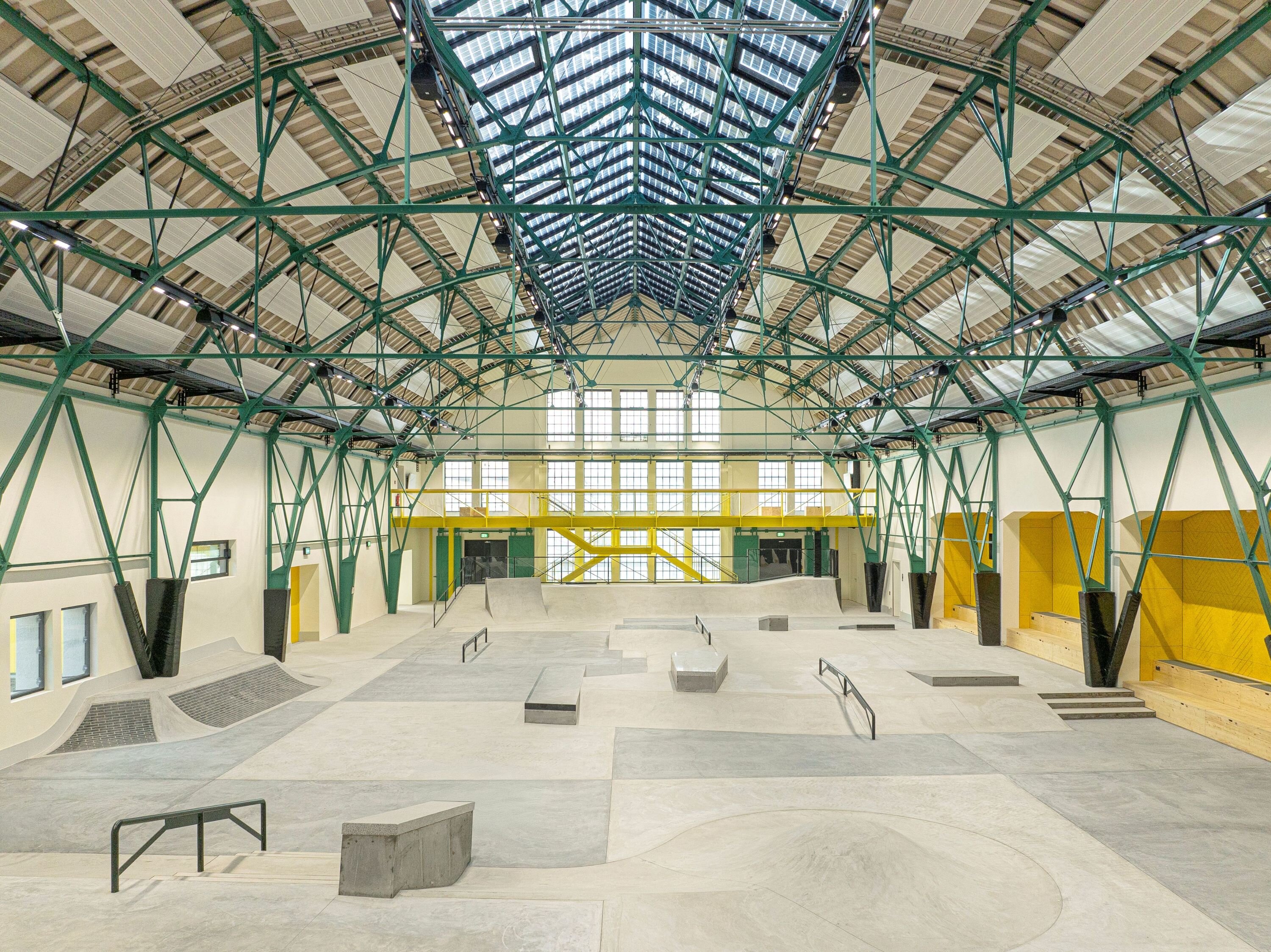 Project by Behnisch Architekturbuero, Action Sport Center. Indoor skatepark with ramps and rails in a large hall with steel truss roof. Yellow and green accents are inside the hall.