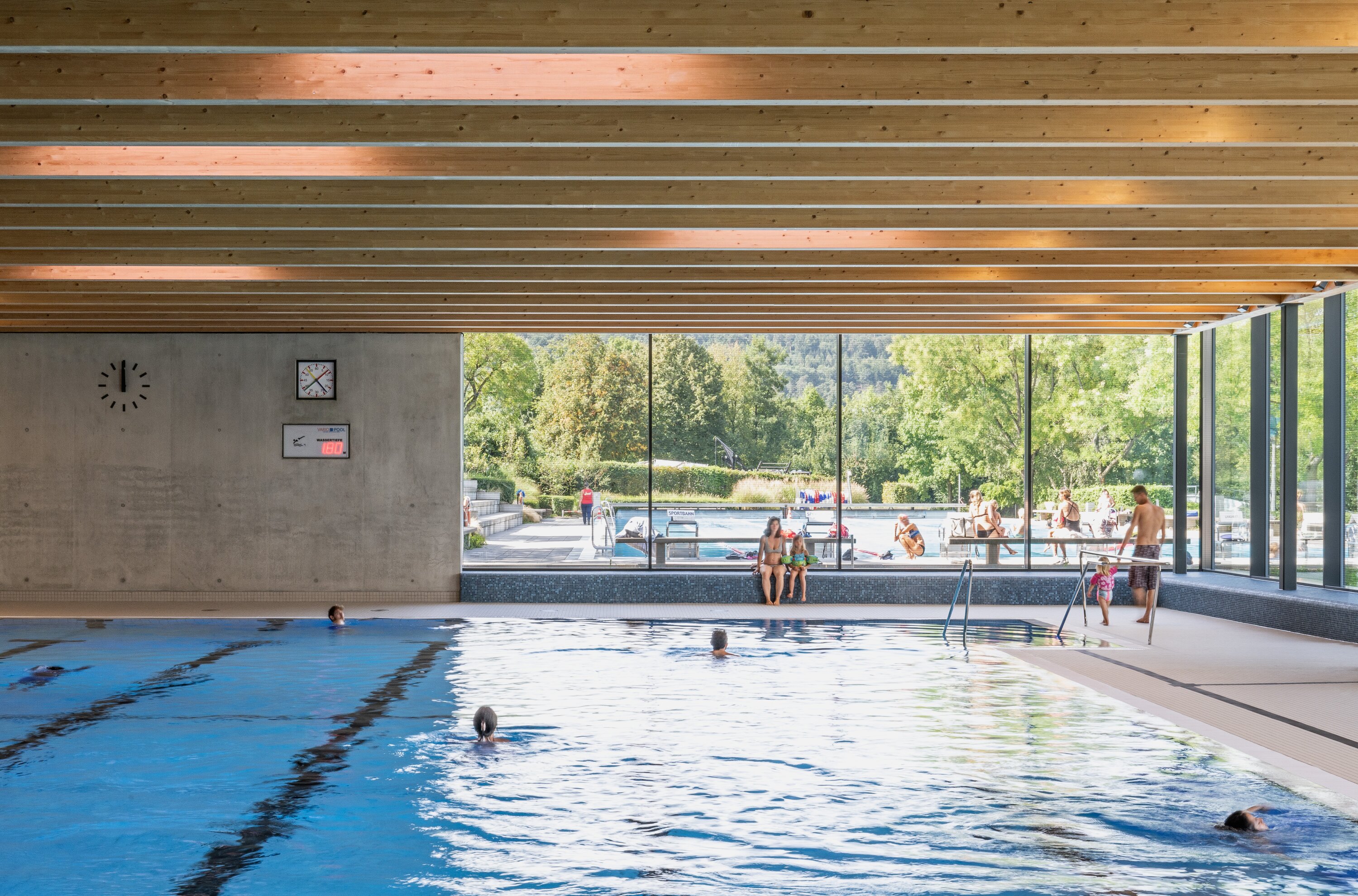 Project by Behnisch Architekturbuero, Wunnebad Winnenden. Indoor swimming pool with a wooden ceiling, large windows, and people swimming in the pool or sitting next to it.