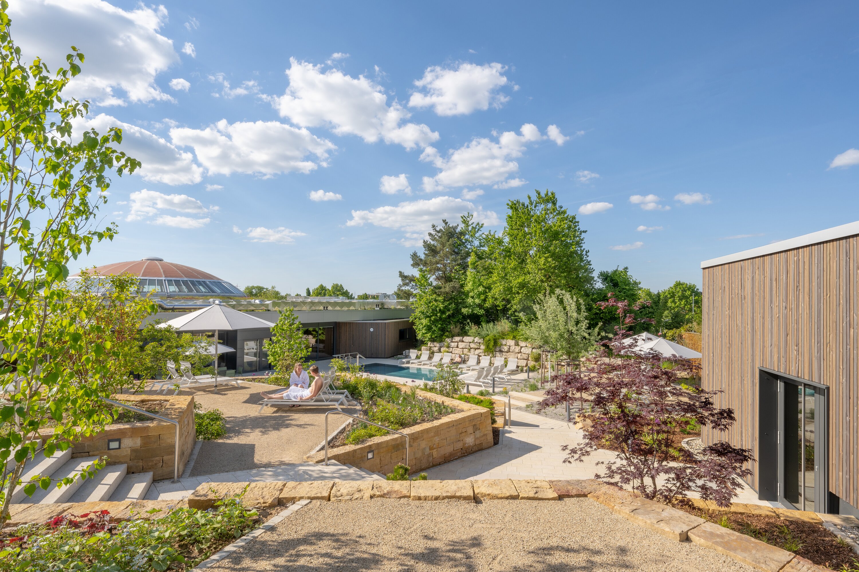 Project by Behnisch Architekturbuero, Wunnebad Winnenden. Landscaped outdoor spa area with pool, lounge chairs, trees, and low wooden buildings.