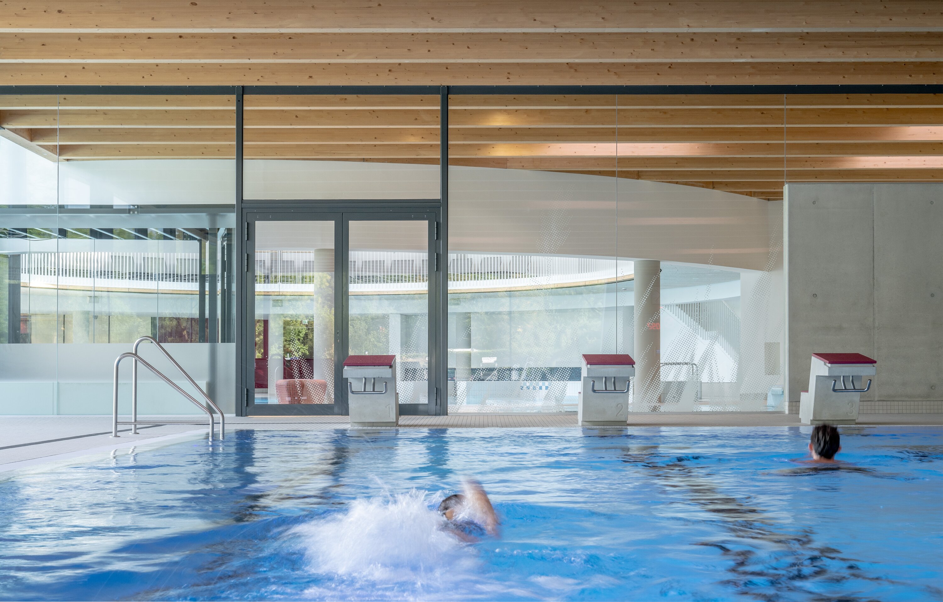 Project by Behnisch Architekturbuero, Wunnebad Winnenden. Two people are swimming in the large pool. Starting blocks can be seen in front of the pool.