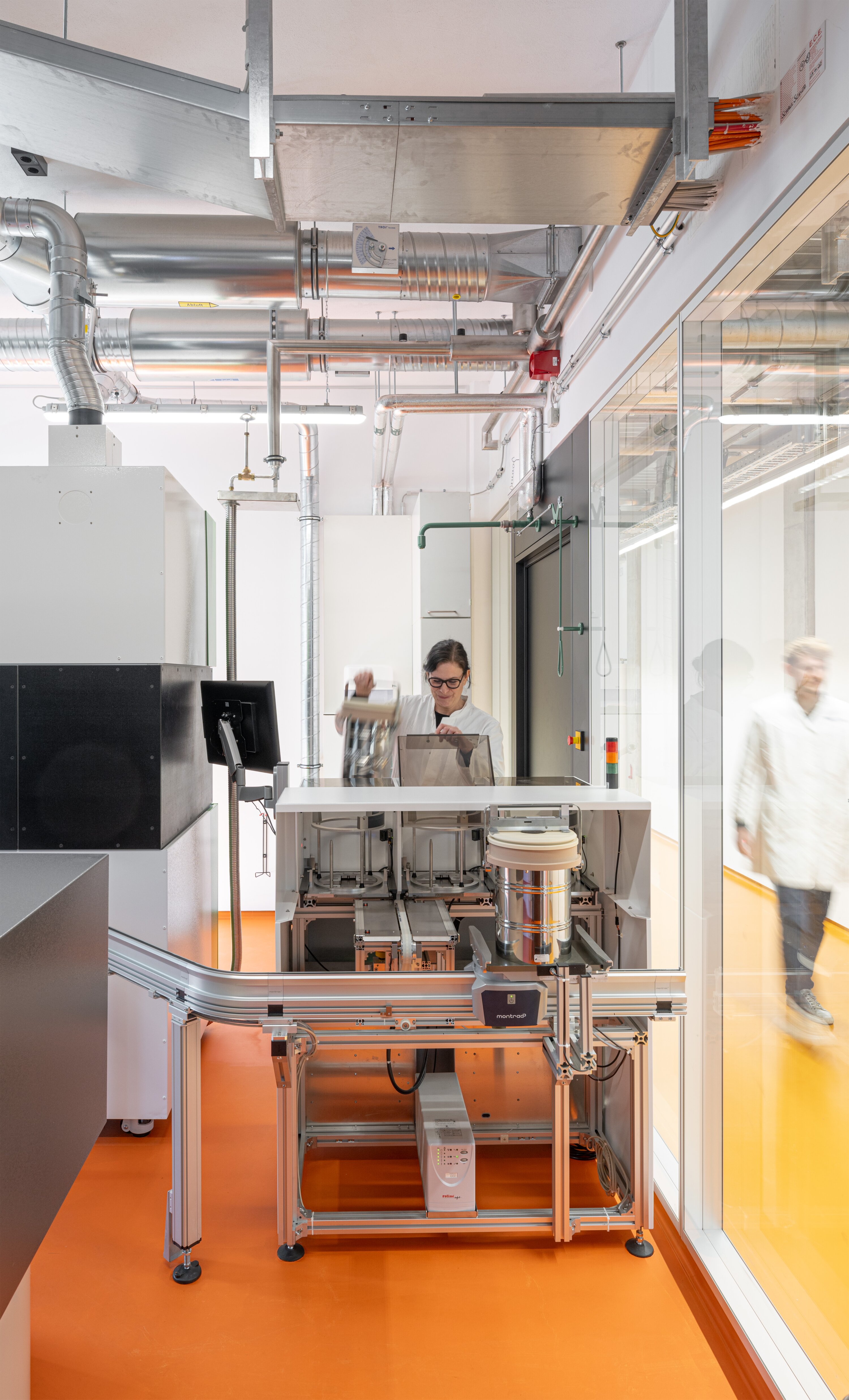 Project by Behnisch Architekturbuero, Dieter Morszeck Biorepository. A technician operates laboratory equipment in a bright laboratory with an orange floor, glass walls, and exposed ventilation ducts on the ceiling. Through the glass wall, a man can be seen walking down the corridor to the right.