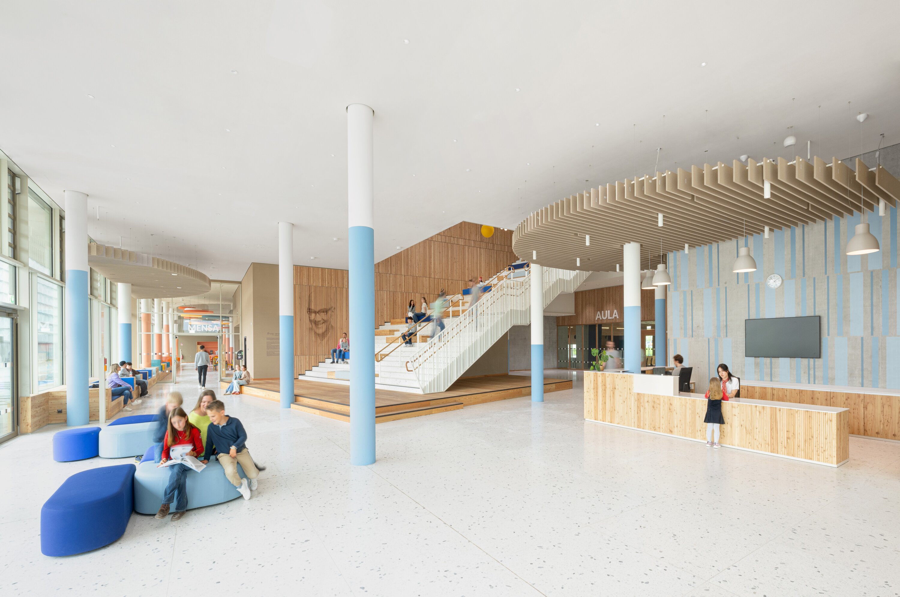 Project by Behnisch Architekturbuero, Josef-Schwarz School. The students gather in a bright school hall with stairs, seating, a reception area, and large windows. Children sit on the dark and light blue chairs and read.