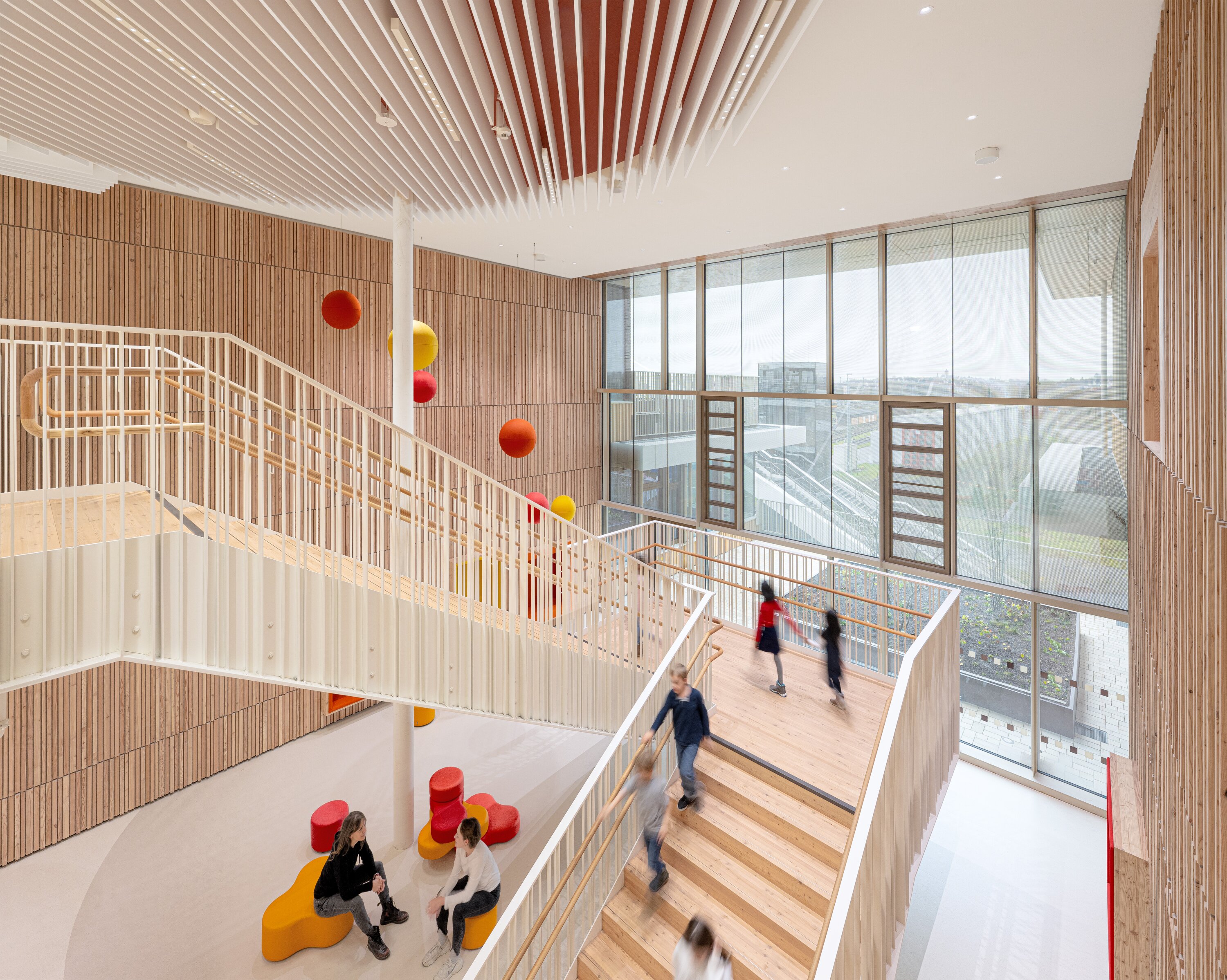 Project by Behnisch Architekturbuero, Josef-Schwarz School. Children move about on the stairs in a bright atrium with wood-paneled walls and large windows. Students sit on the colorful seating on the lower floor. Colorful spheres hang from the ceiling.