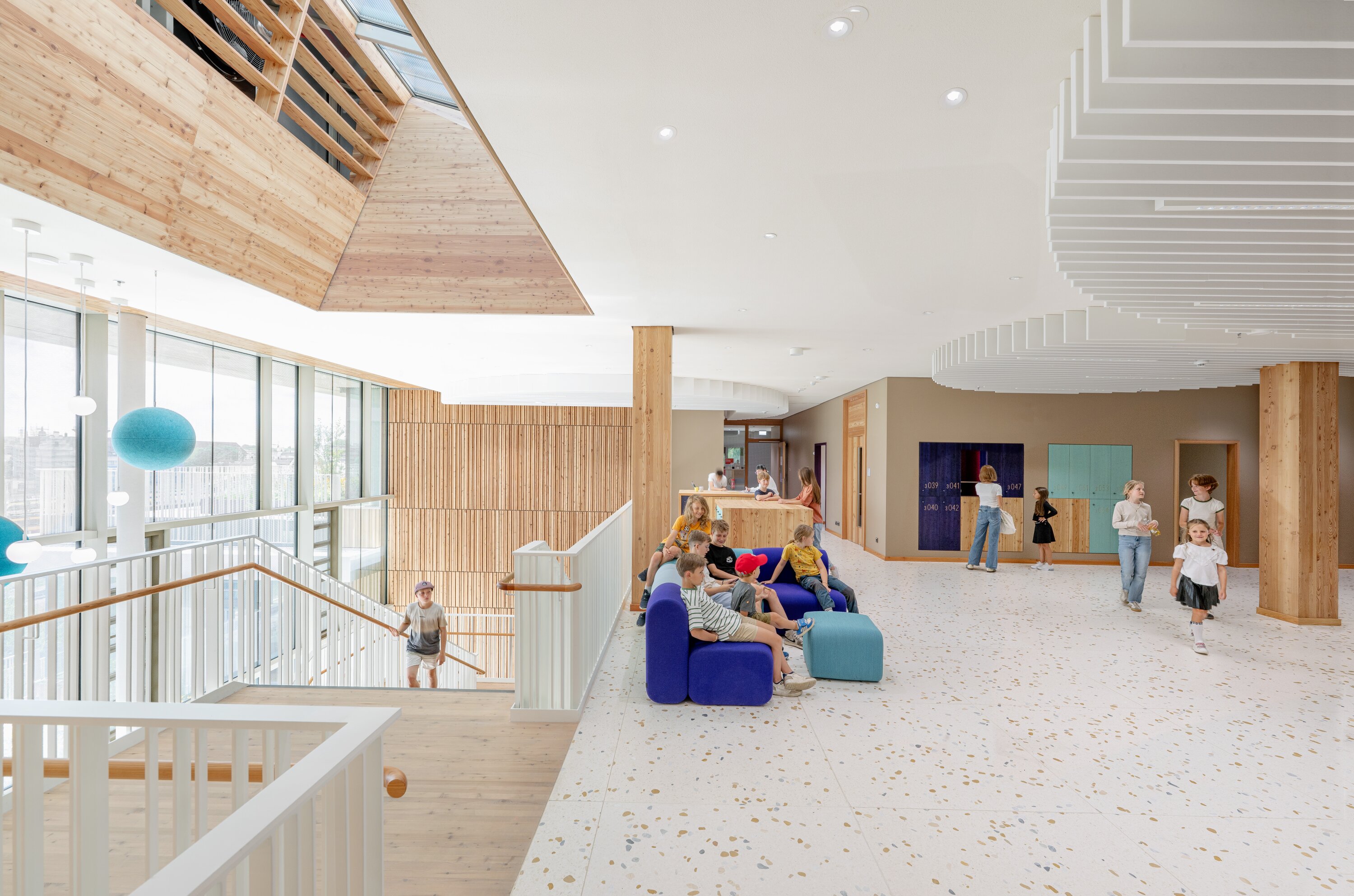 Project by Behnisch Architekturbuero, Josef-Schwarz School. Children and adults in a bright school lobby with stairs, seating, and wooden finishes.