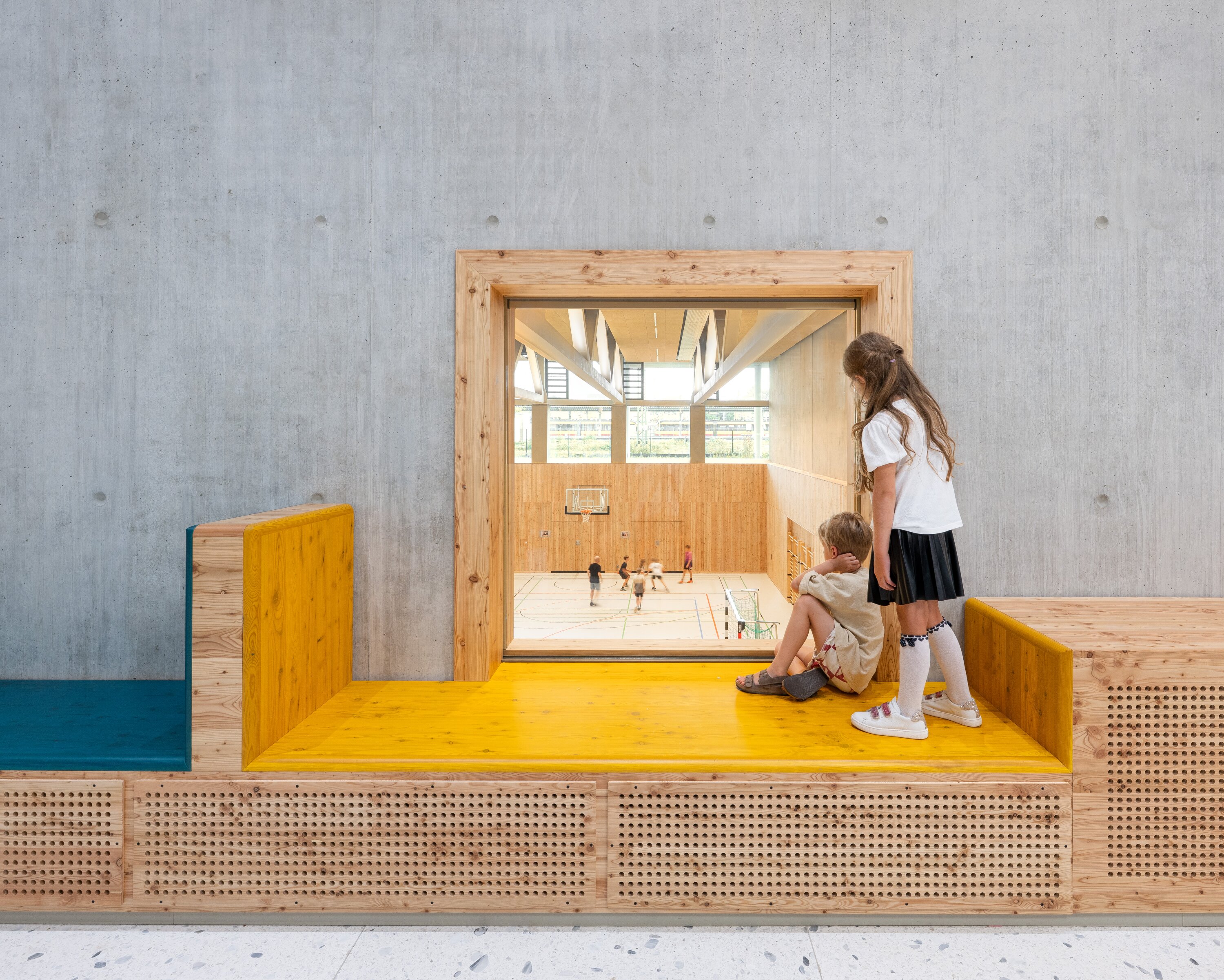 Project by Behnisch Architekturbuero, Josef-Schwarz School. Two children are sitting on a wooden bench by a window overlooking the sports hall. Inside the hall, other children are playing basketball.