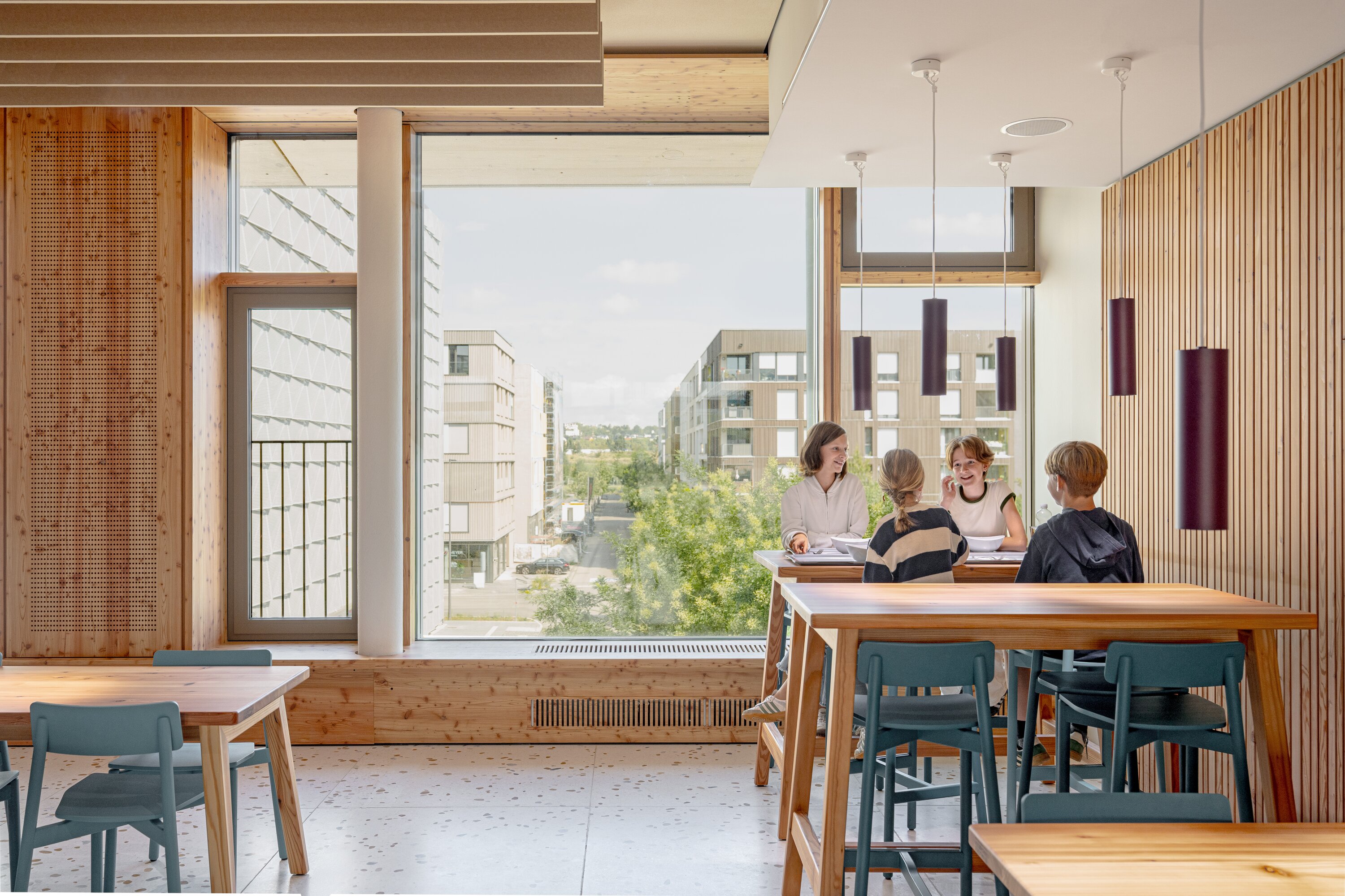 Project by Behnisch Architekturbuero, Josef-Schwarz School. Children and an adult sit at a high table in a bright room with large window overlooking buildings.