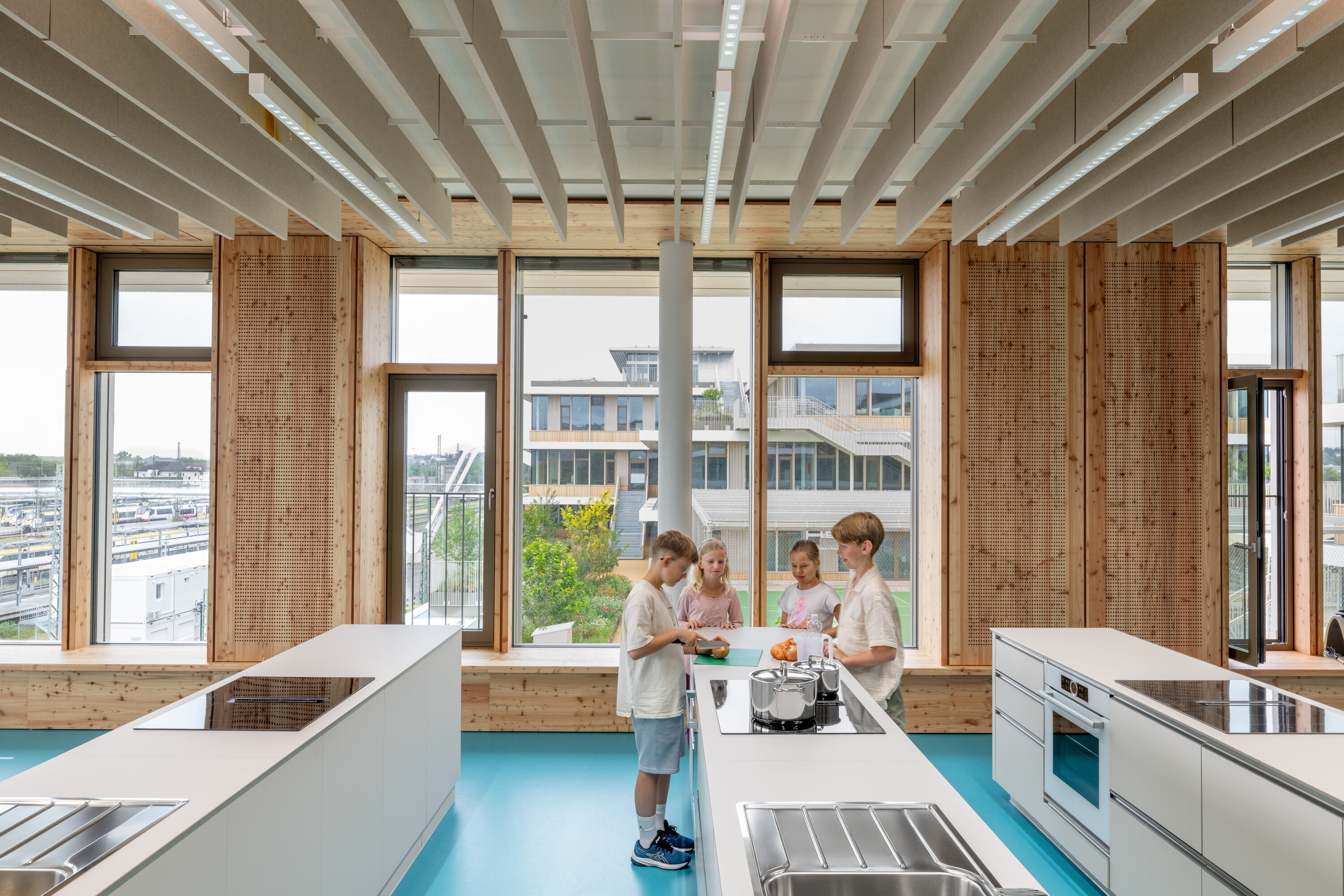Project by Behnisch Architekturbuero, Josef-Schwarz School. In a bright room with large windows, children are preparing food at one of the three kitchen islands. Through the generous window front, the outdoor area with a football field is visible in the background.