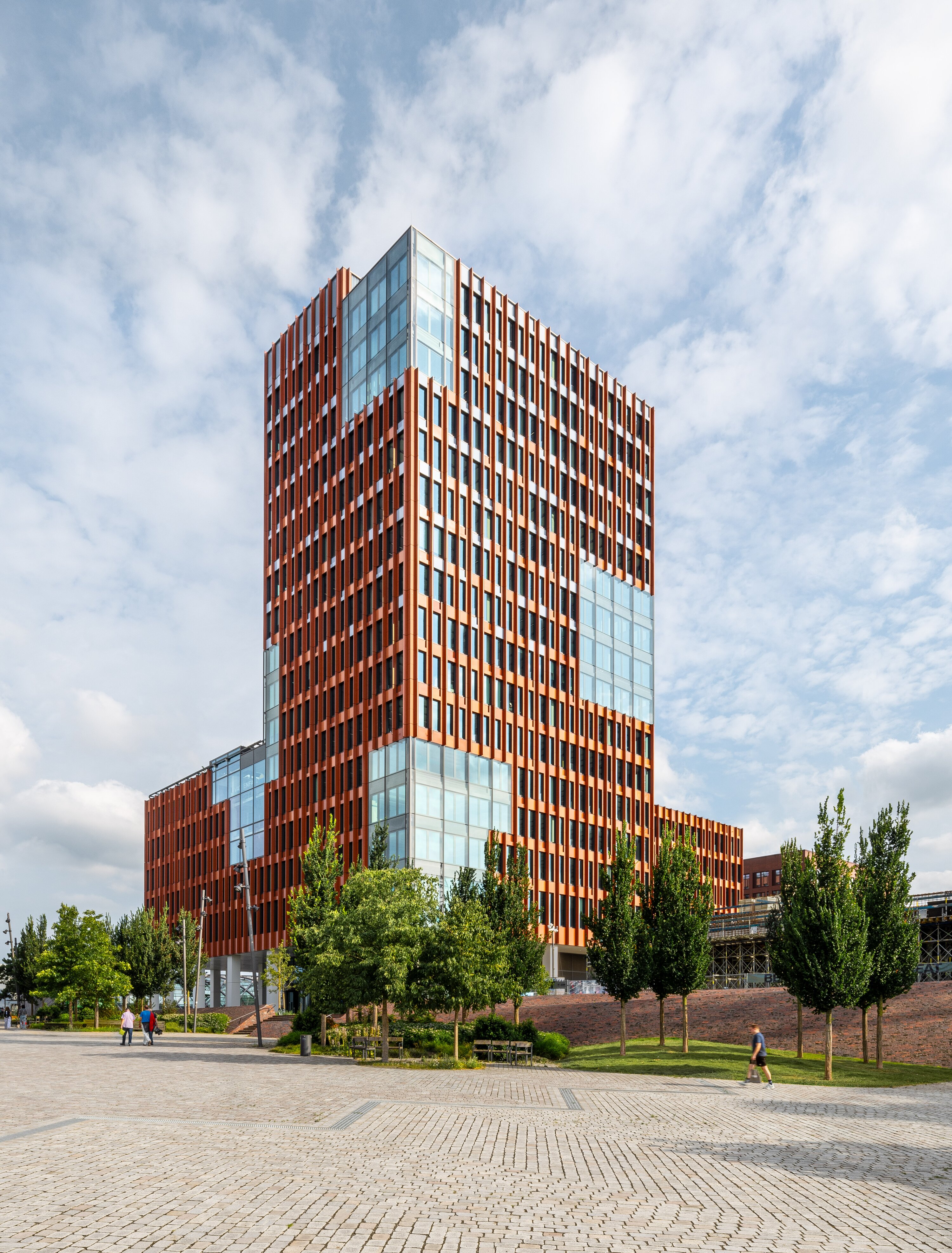 Project by Behnisch Architekturbuero, EDGE ElbSide Hamburg. Red high-rise building with glass sections, trees and pedestrians in foreground.