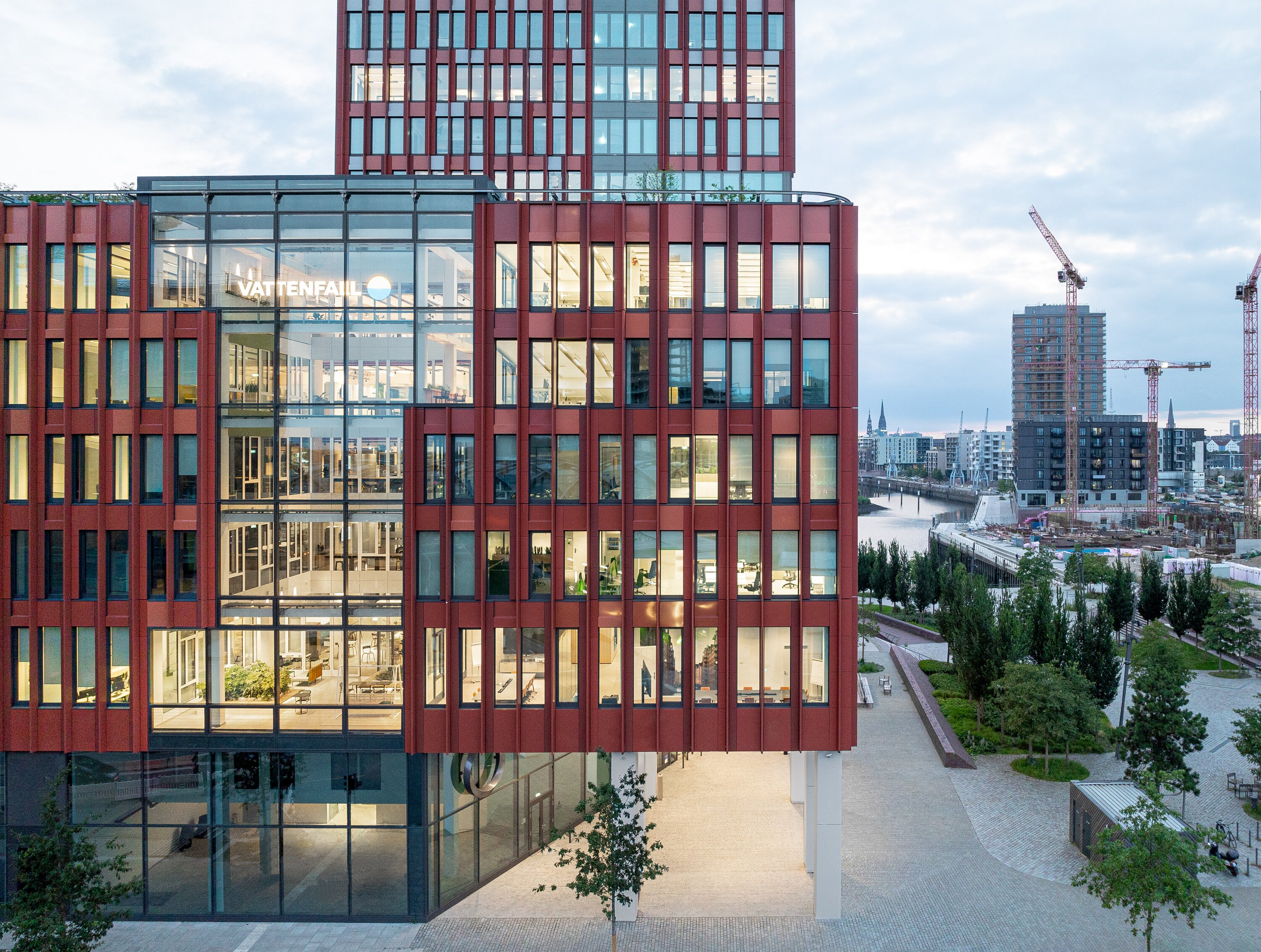 Project by Behnisch Architekturbuero, EDGE ElbSide Hamburg. Office building with a reddish facade. View into the glass atrium and the office spaces. In the background, the river and the surrounding city can be seen. The green forecourt is visible on the right.