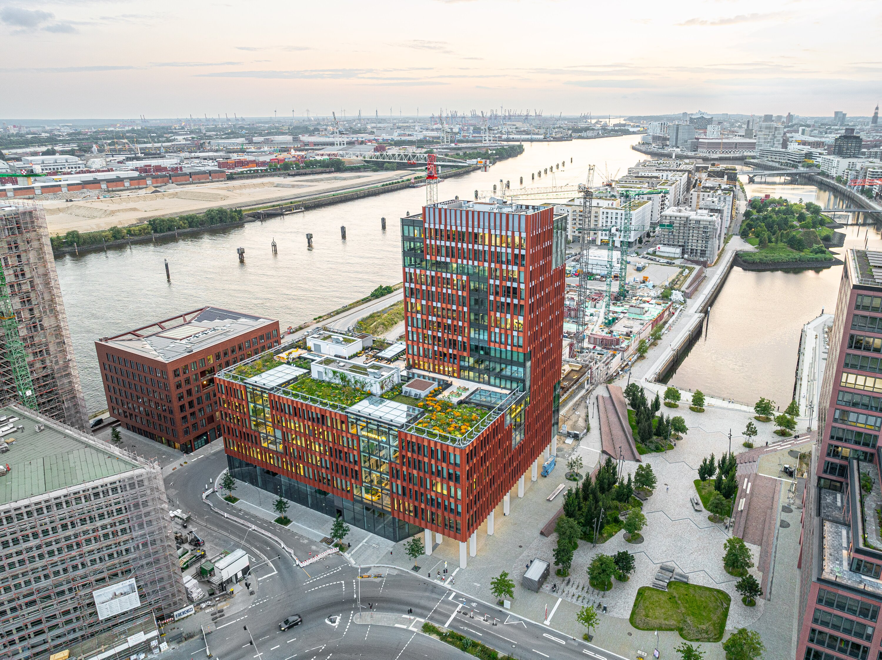Project by Behnisch Architekturbuero, EDGE ElbSide Hamburg. Aerial view of a red-facing office building overlooking the river and the surrounding city. To the right of the building is a public square with seating, bicycle racks, green spaces, and trees.