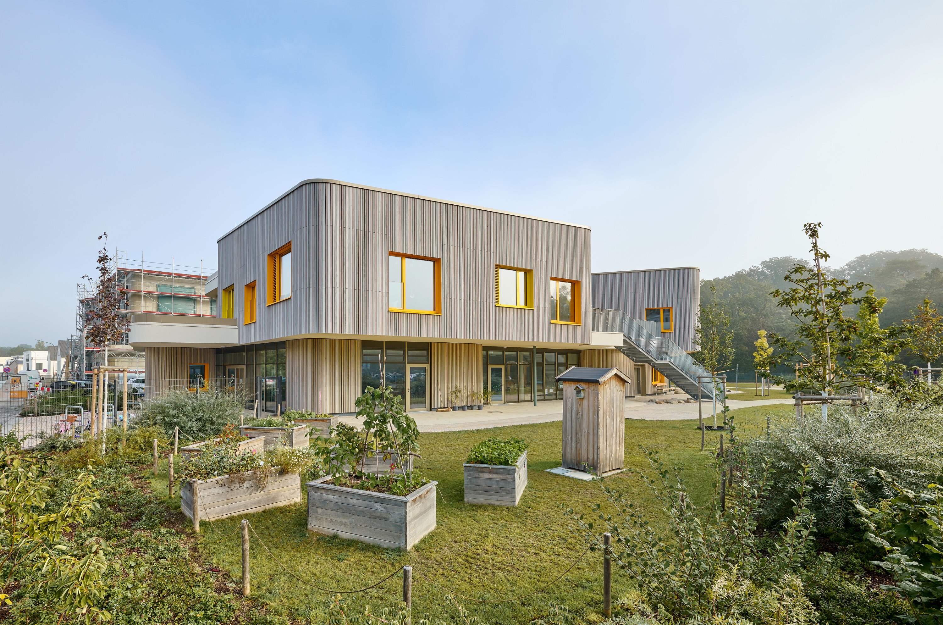 Project by Behnisch Architekturbuero, KiTa Pioneer-Kaserne. Modern wooden building with yellow and orange framed windows. Surrounded by a garden with raised beds and lawns.