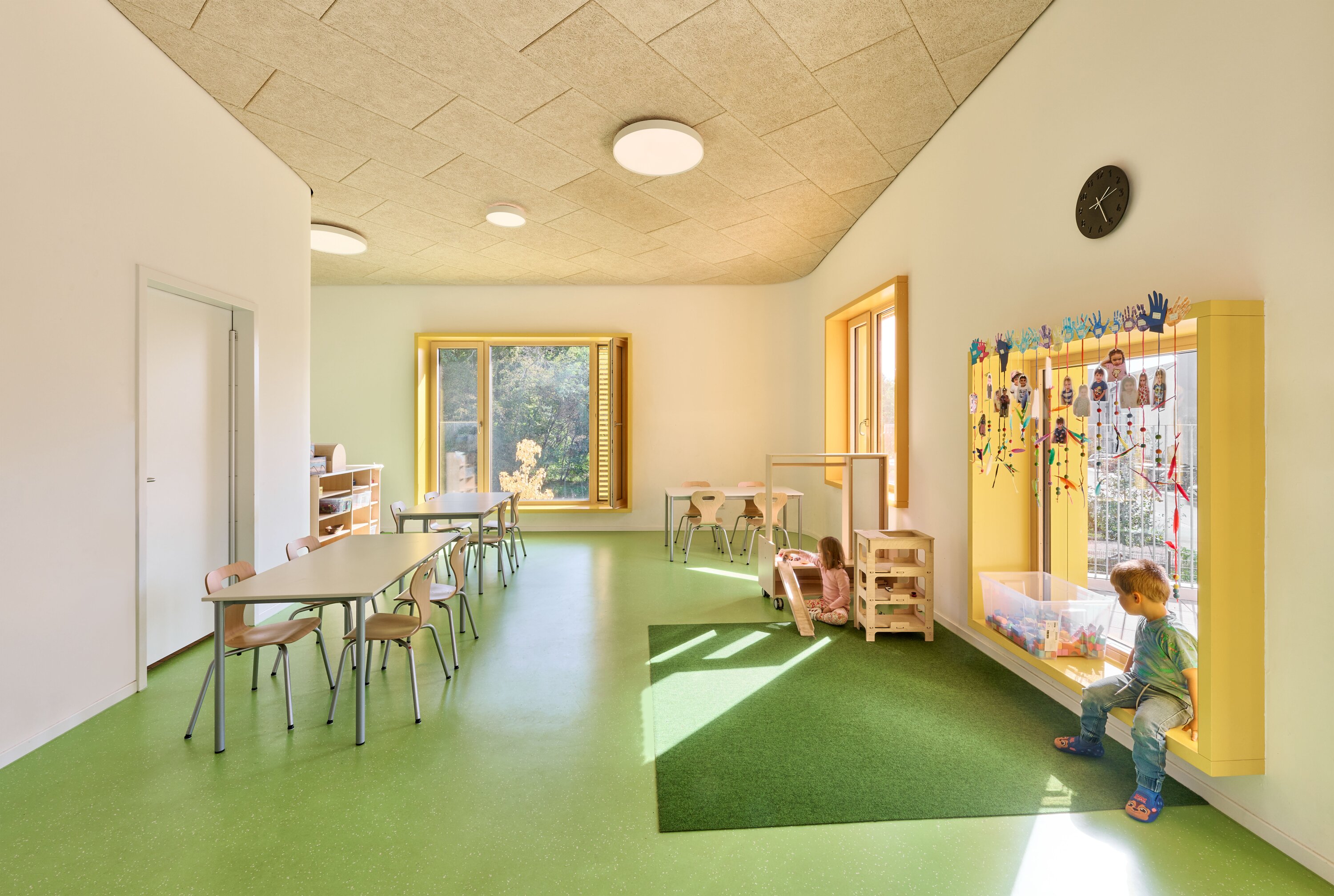 Project by Behnisch Architekturbuero, KiTa Pioneer-Kaserne. Bright playroom with green floor, tables and chairs, large windows and children playing near a yellow windowsill and a toy storage room.